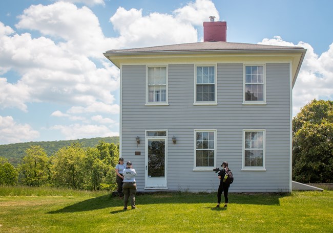 Three employees in front of a two-story blue historic house. Two of the employees are wearing NPS uniforms and the other one is wearing an intern uniform. One of the NPS workers is talking while the other worker and intern are filming.