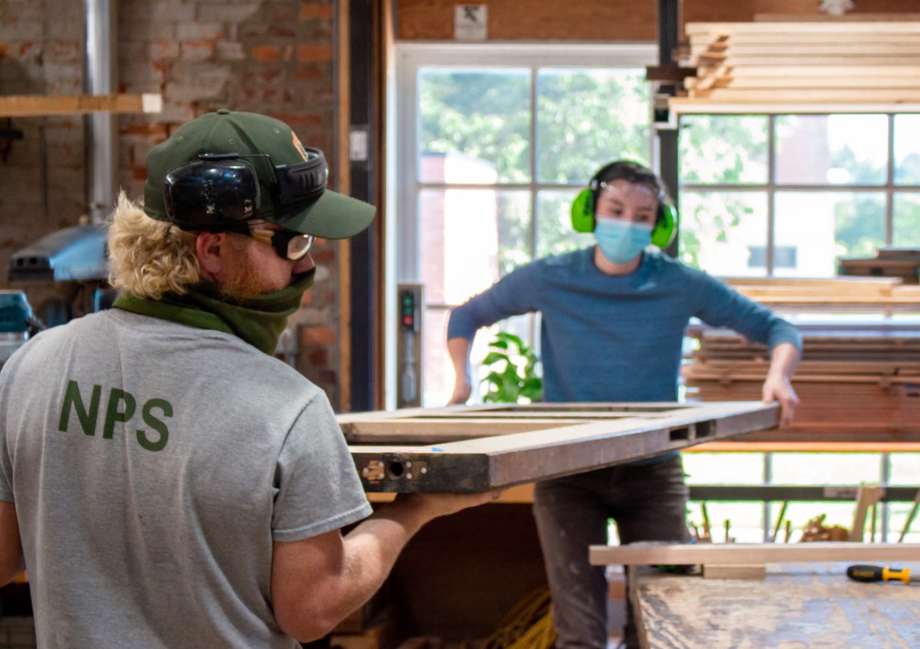 Two people carry a heavy door inside a workshop.