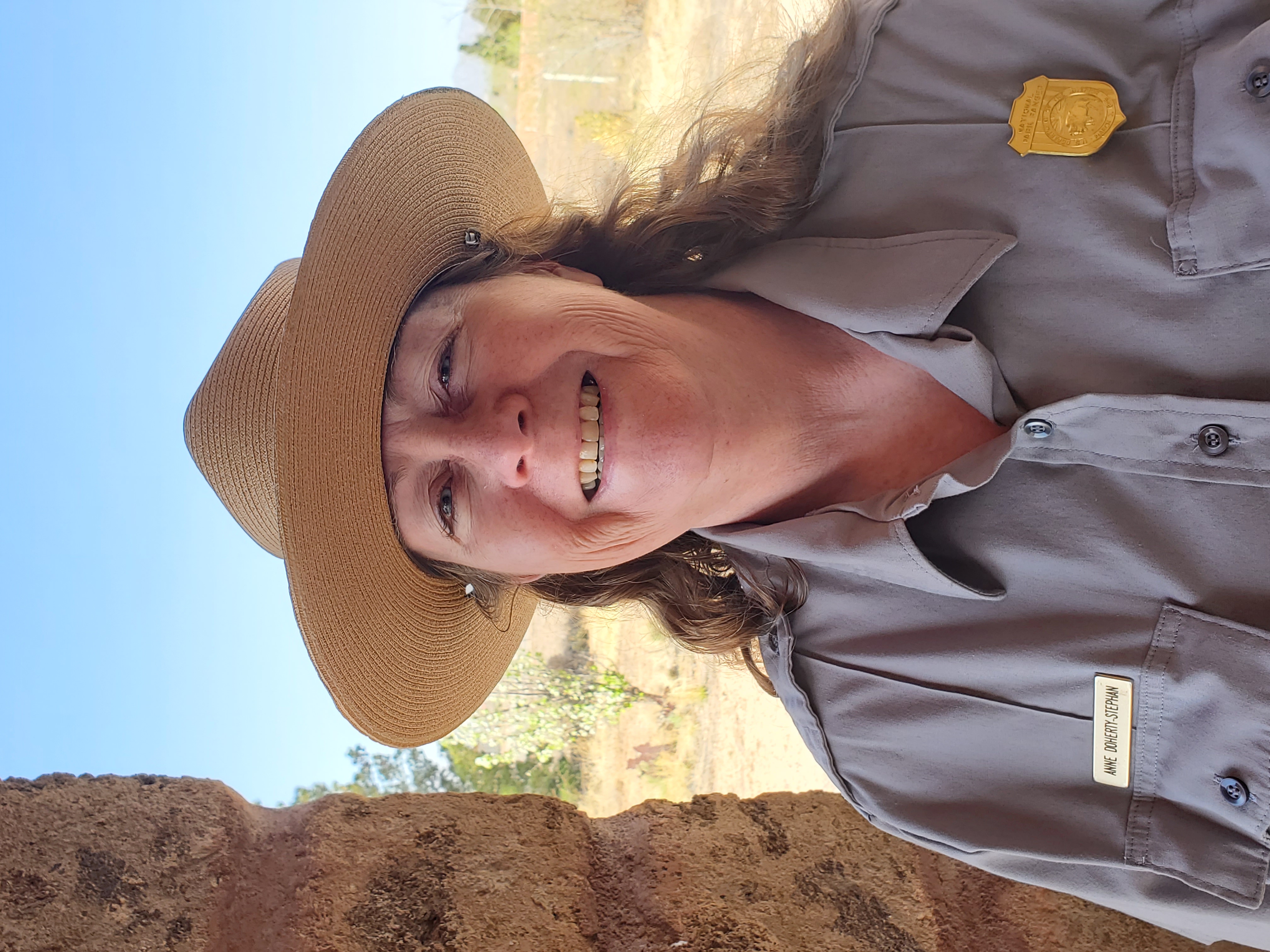 Anne Doherty-Stephan selected as superintendent of César E. Chávez National Monument Woman in NPS uniform and flat hat stands beside stone pillar. An arid background with sparse trees and blue skies are behind her.