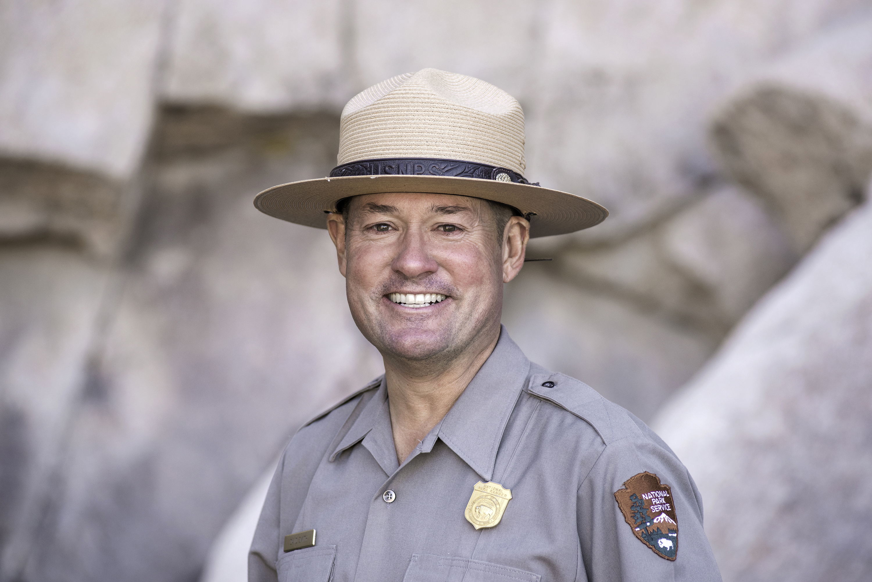 A white man smiles in a gray NPS uniform shirt and tan flat hat stands in front of a tan/gray rock wall. He is only visable from the chest up.
