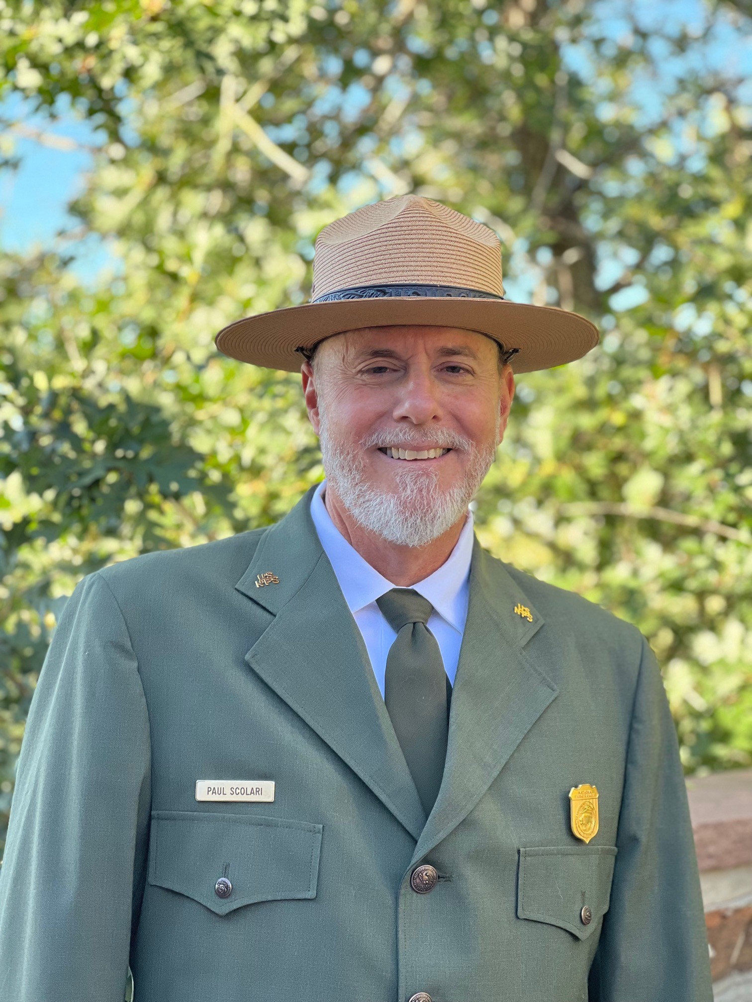 Scolari KAHO-Puho Bio Pic Portrait of a man with a white beard in formal NPS uniform standing in front of green foliage.