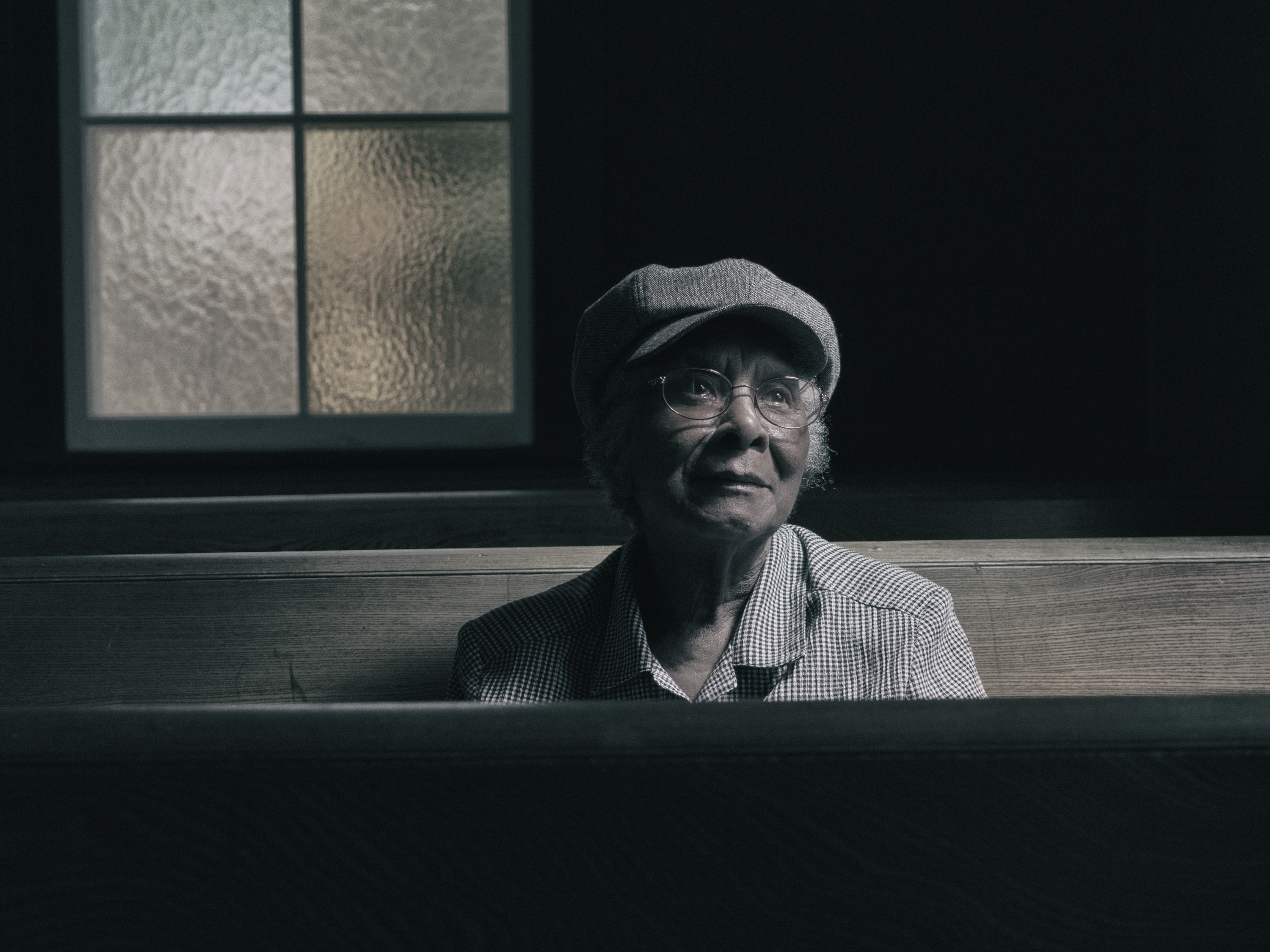 A woman sits within a wooden pew gazing up and to the right. She sits in a dark space with light shining over her face. A glass window is behind her right shoulder. She wears glasses, a gray hat, a checkered shirt, and has gazes softly into the distance.