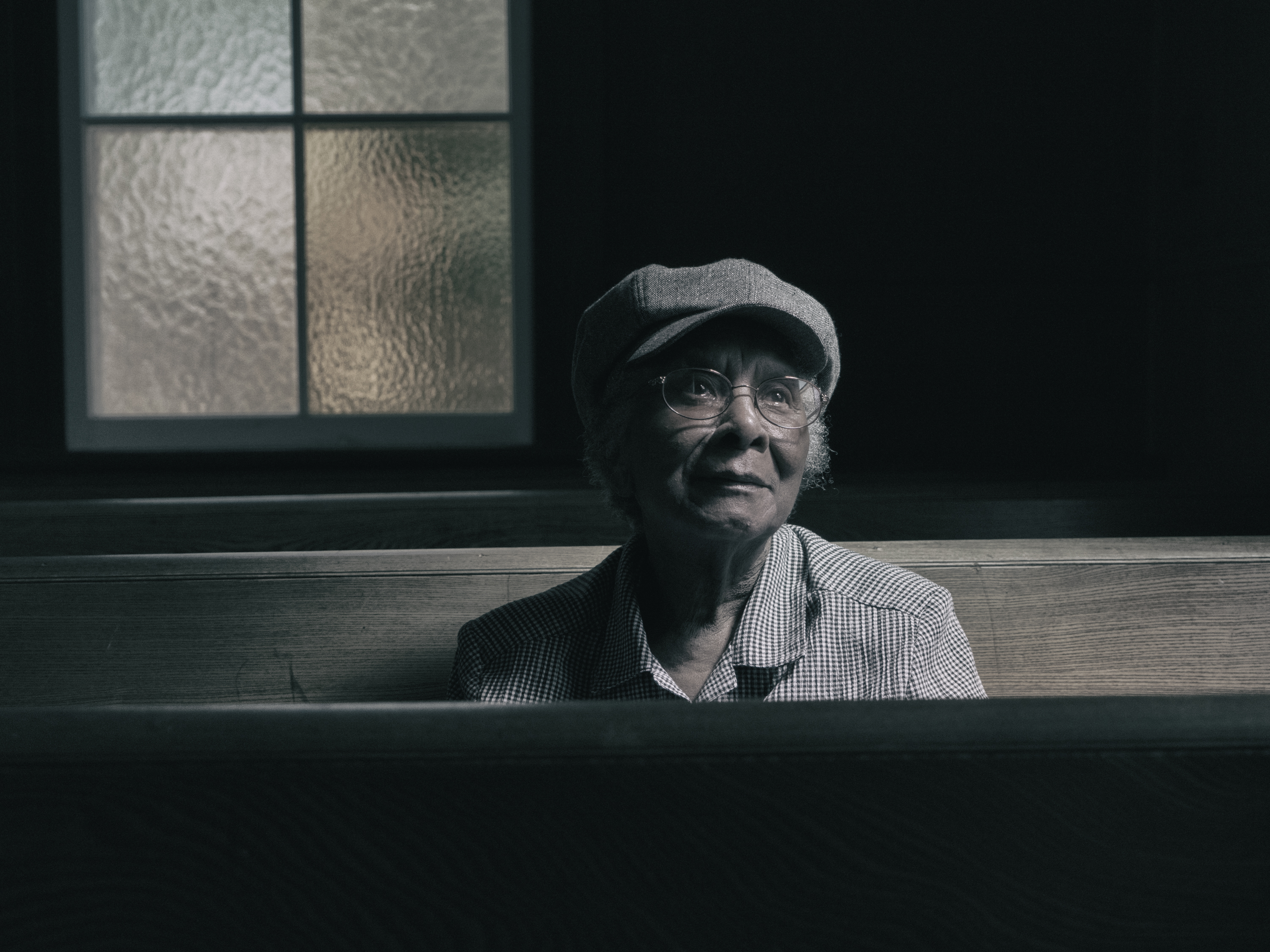 All photographs are owned by Yuri Dojc and protected by copyright law. To reproduce or distribute these contents without written permission of the owner is prohibited. A woman sits within a wooden pew gazing up and to the right. She sits in a dark space with light shining over her face. A glass window is behind her right shoulder. She wears glasses, a gray hat, a checkered shirt, and has gazes softly into the distance.