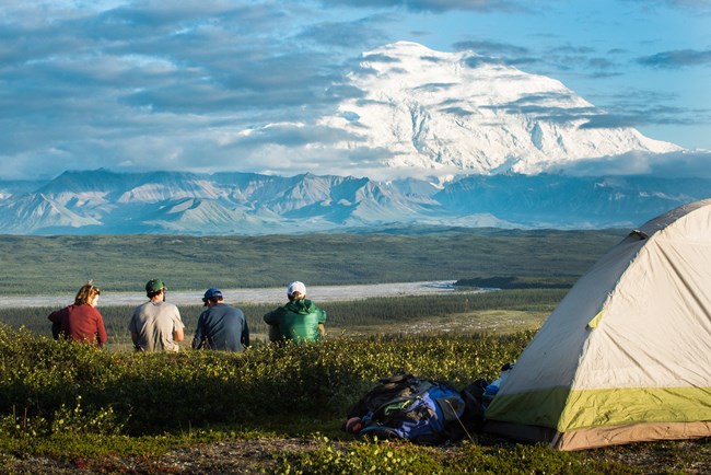 Four people sit by a tent in a grassy field that overlooks a valley, with a large mountain in the distance.