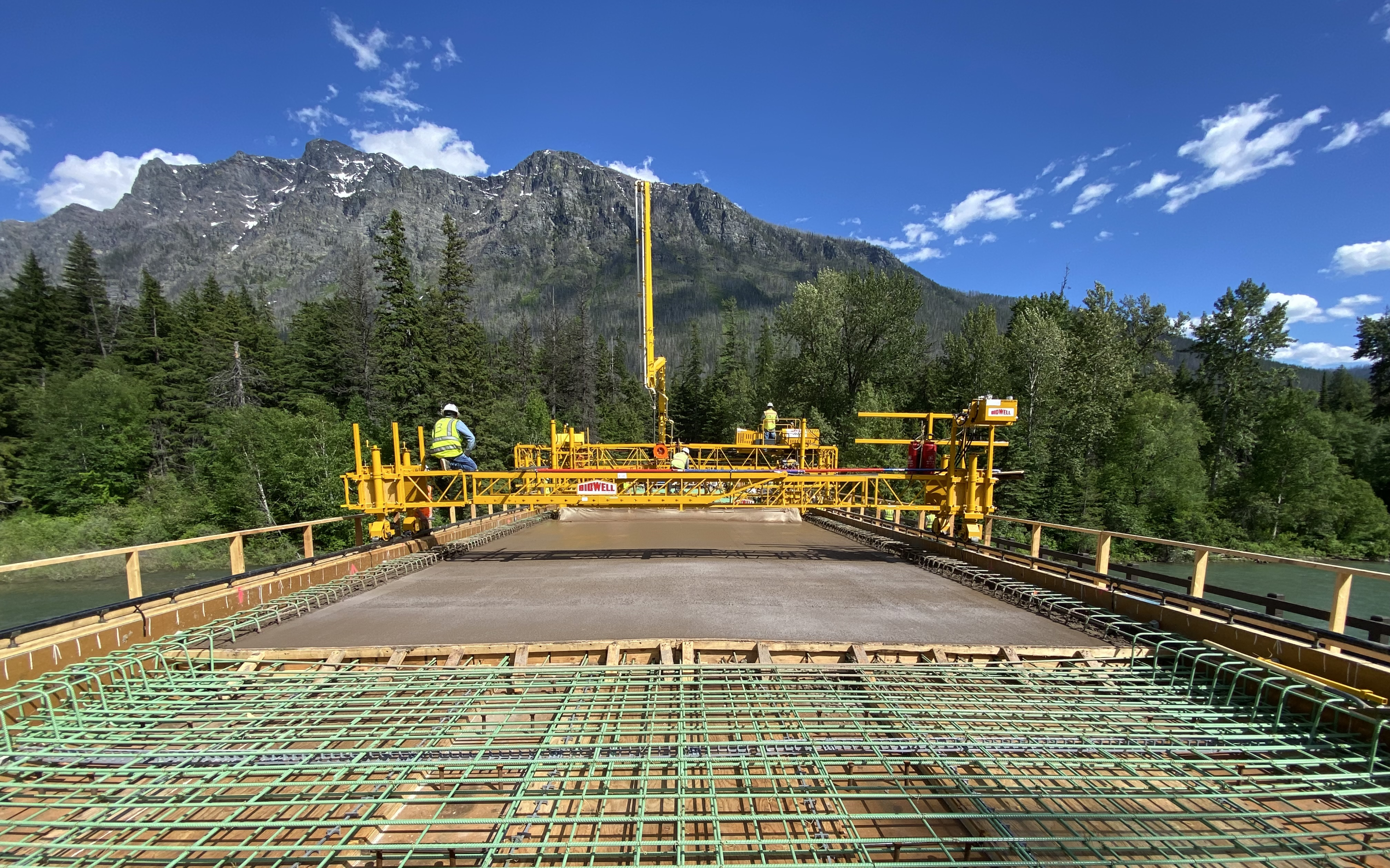 Against a backdrop of mountains and pine trees, a bridge is being constructed.