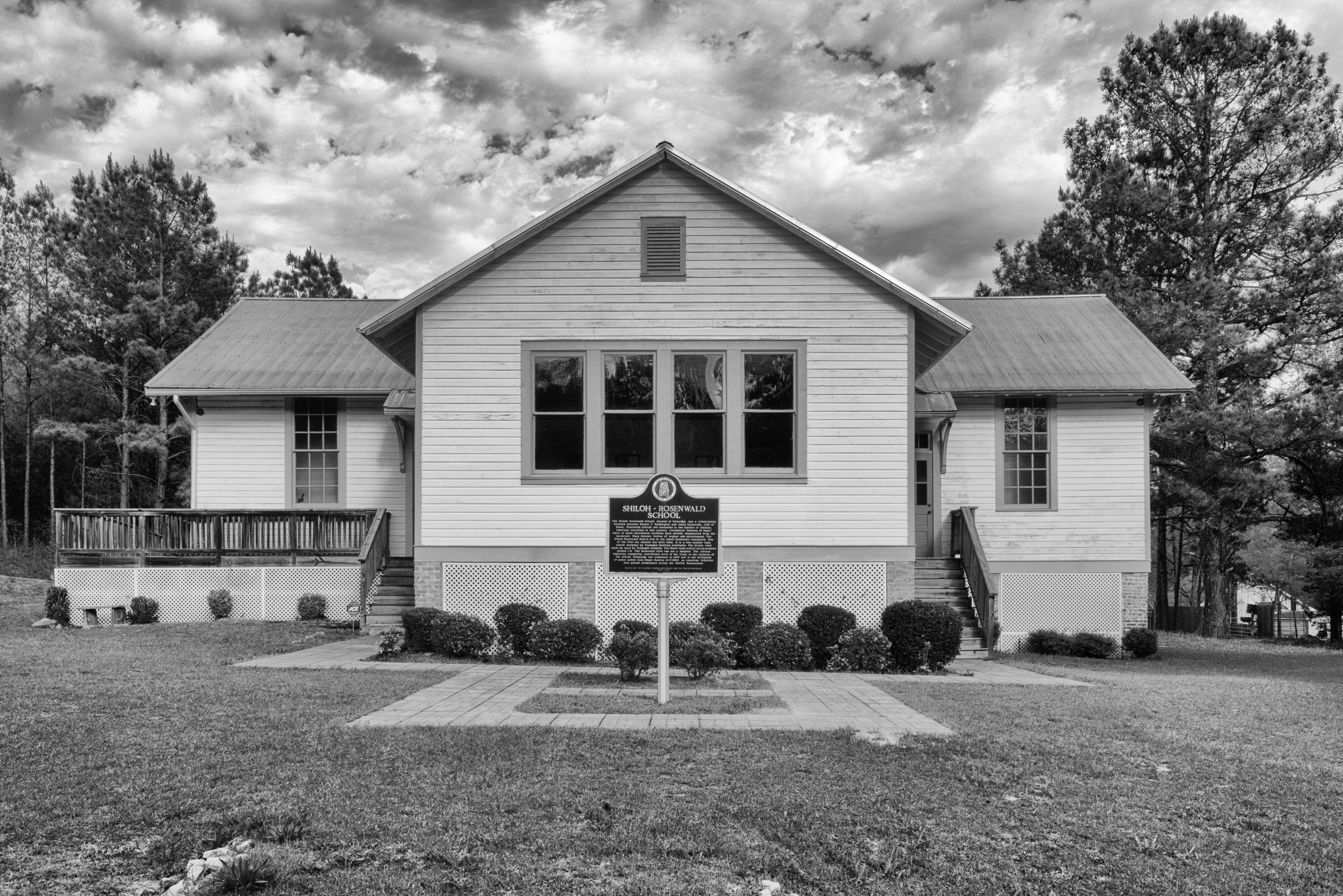 A black and white photo of a small, one-story school surrounded by trees. A sign stands in front of the school that says, "Shiloh-Rosenwald School", followed by small font text that isn't legible.