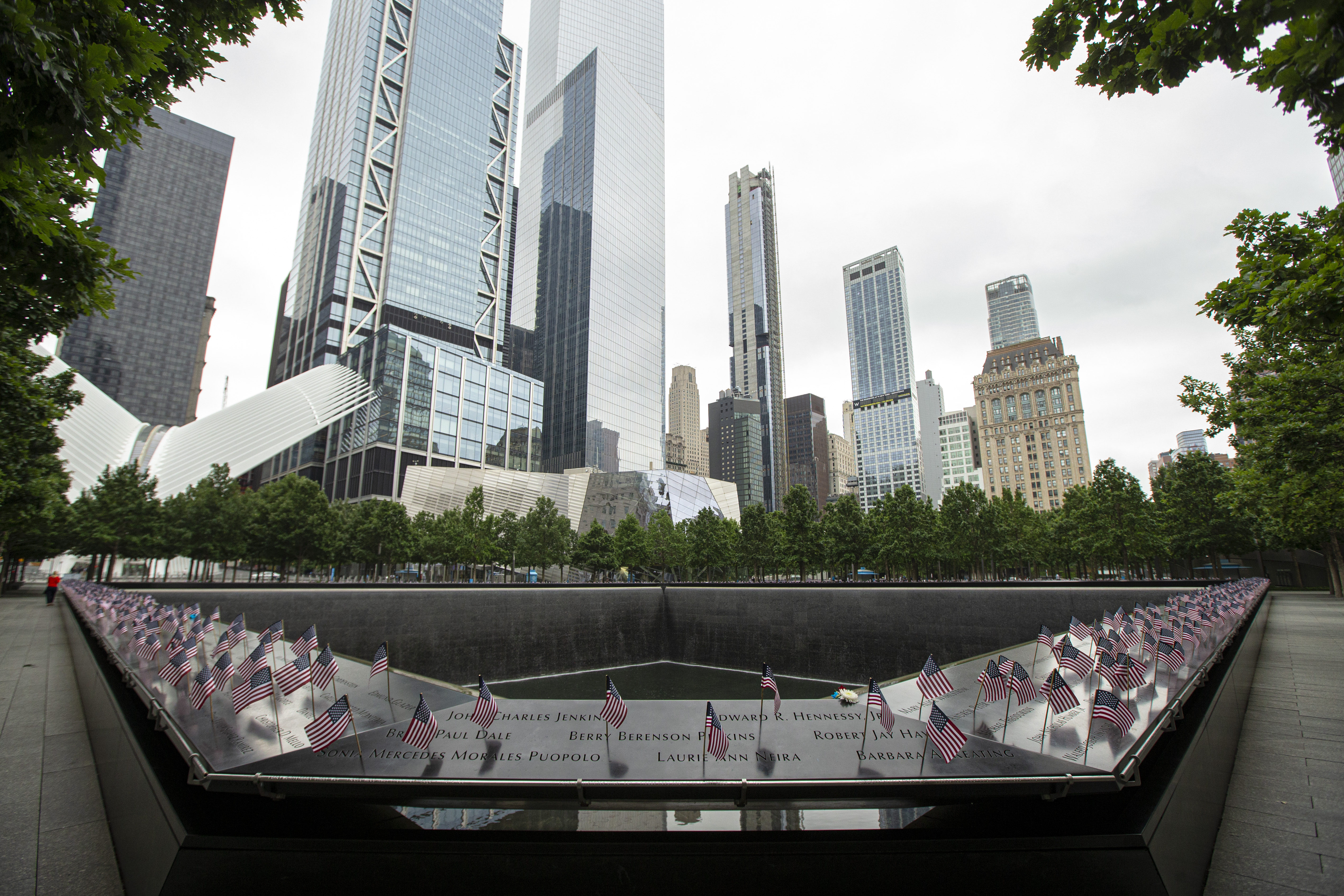 A low, marble square-shaped structure bearing the names of those who perished in the terrorist attacks on 9/11. Small American flags have been placed at each name. In the center of the structure is a deep pool. Tall skyscrapers in the background.
