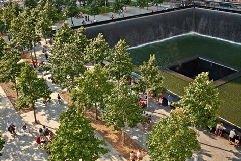 Aerial shot of a large rectangular black memorial with trees in the foreground. The memorial is black, reflective surface built into the ground, with water flowing over the sides down into the pool on the ground.