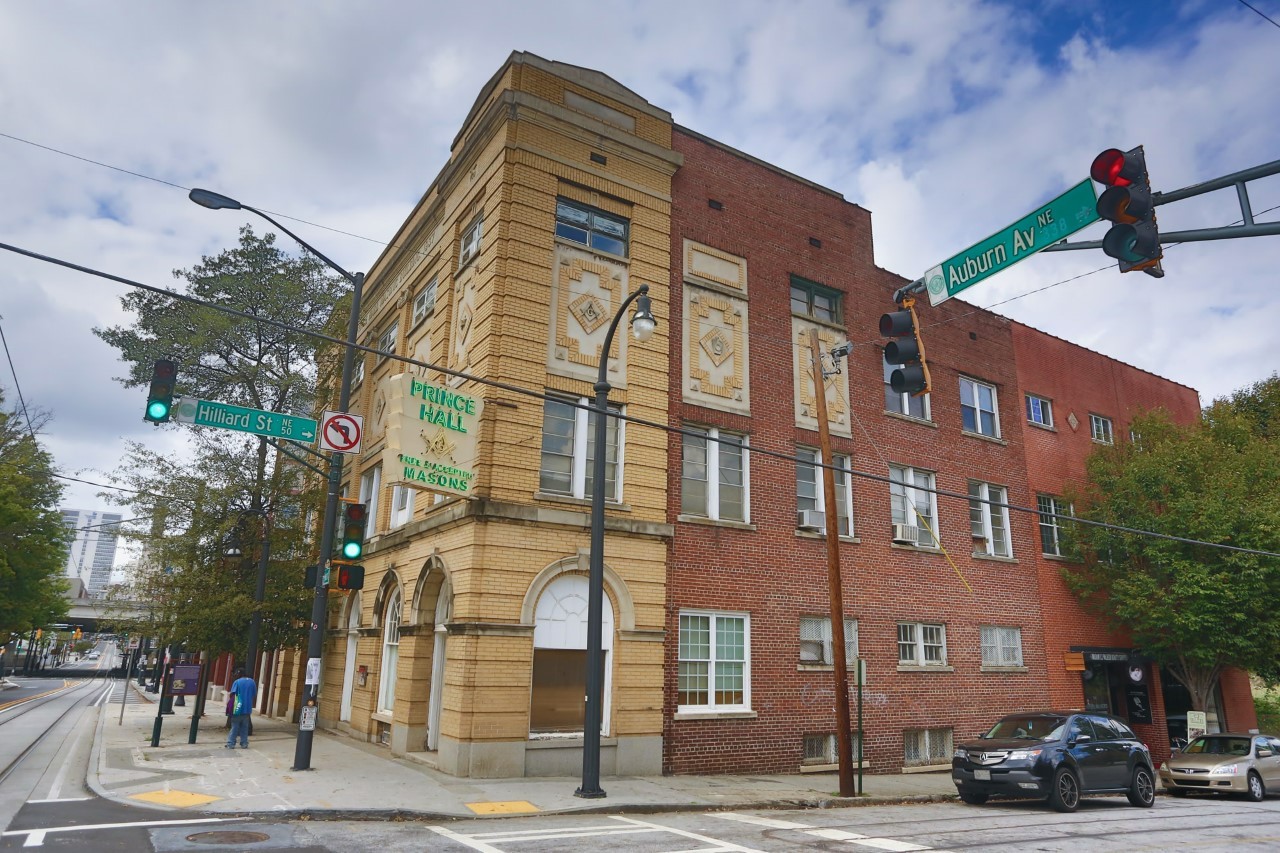 A street corner with a yellow and red brick building with a lit, neon sign that says Prince Hall.