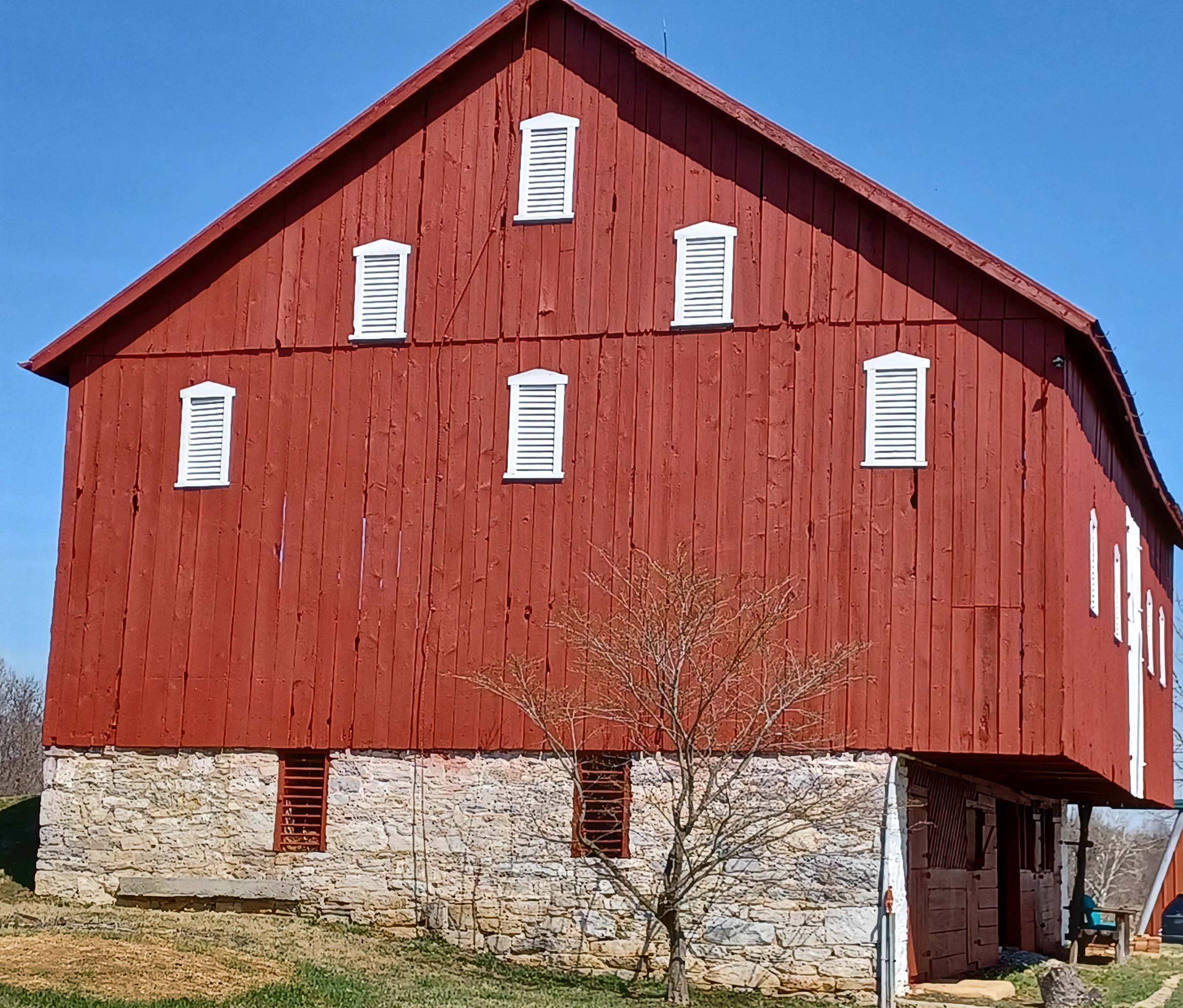 A red, multi-story barn with a stone foundation and white shutters fills the center of the image with a clear blue sky above the building and green glass in the foreground of the building.