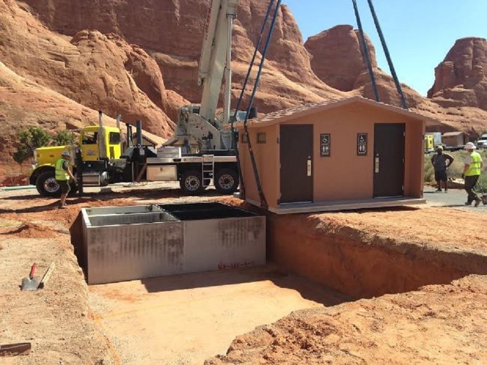 Crane lifting a vault toilet station to move on top of an exposed concrete foundation