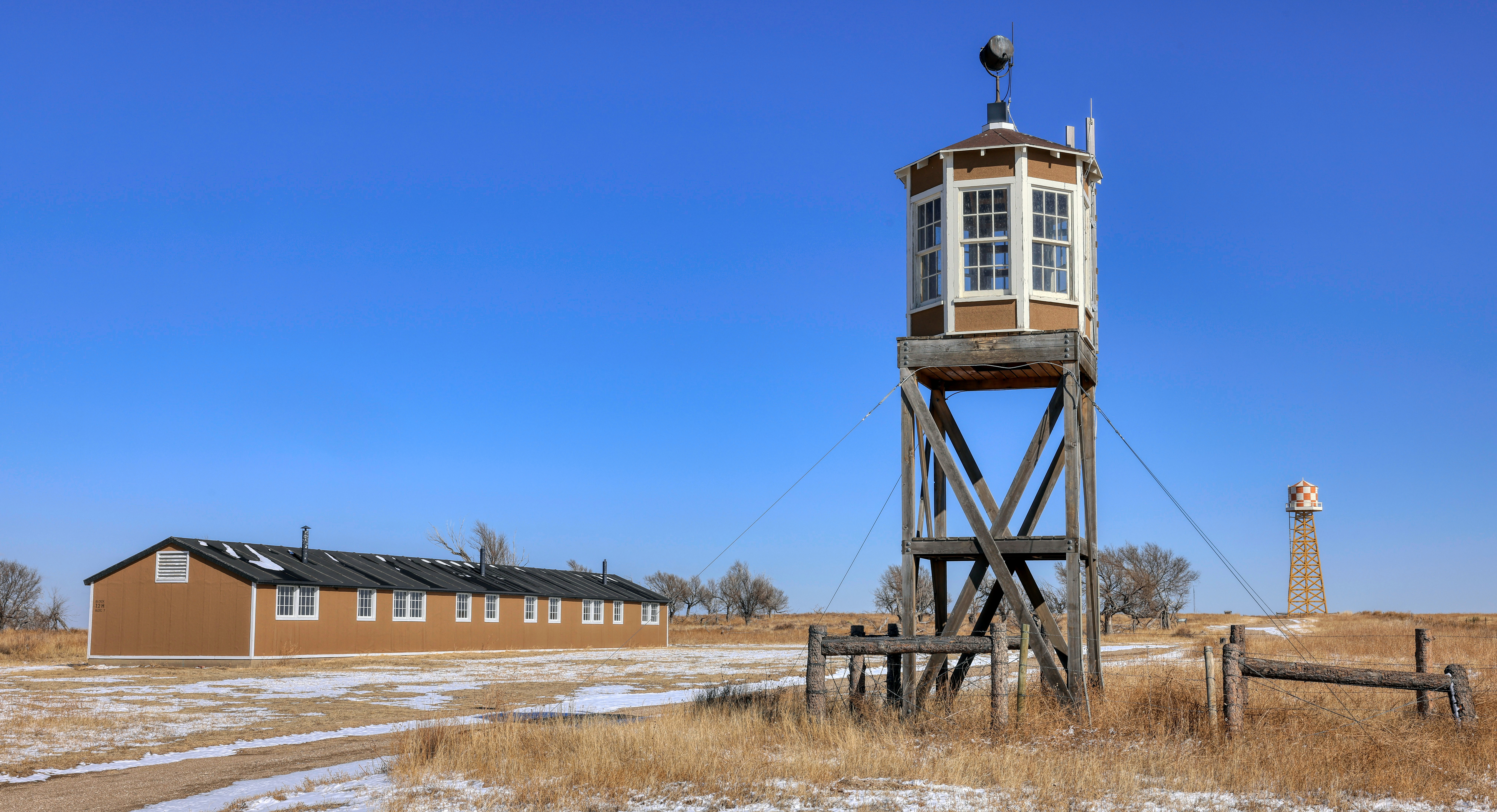 Image of reconstructed barracks nearby a wooden guard tower and fence surrounded by brown grass and shrubs. A reconstructed water tower is visible in the distance.