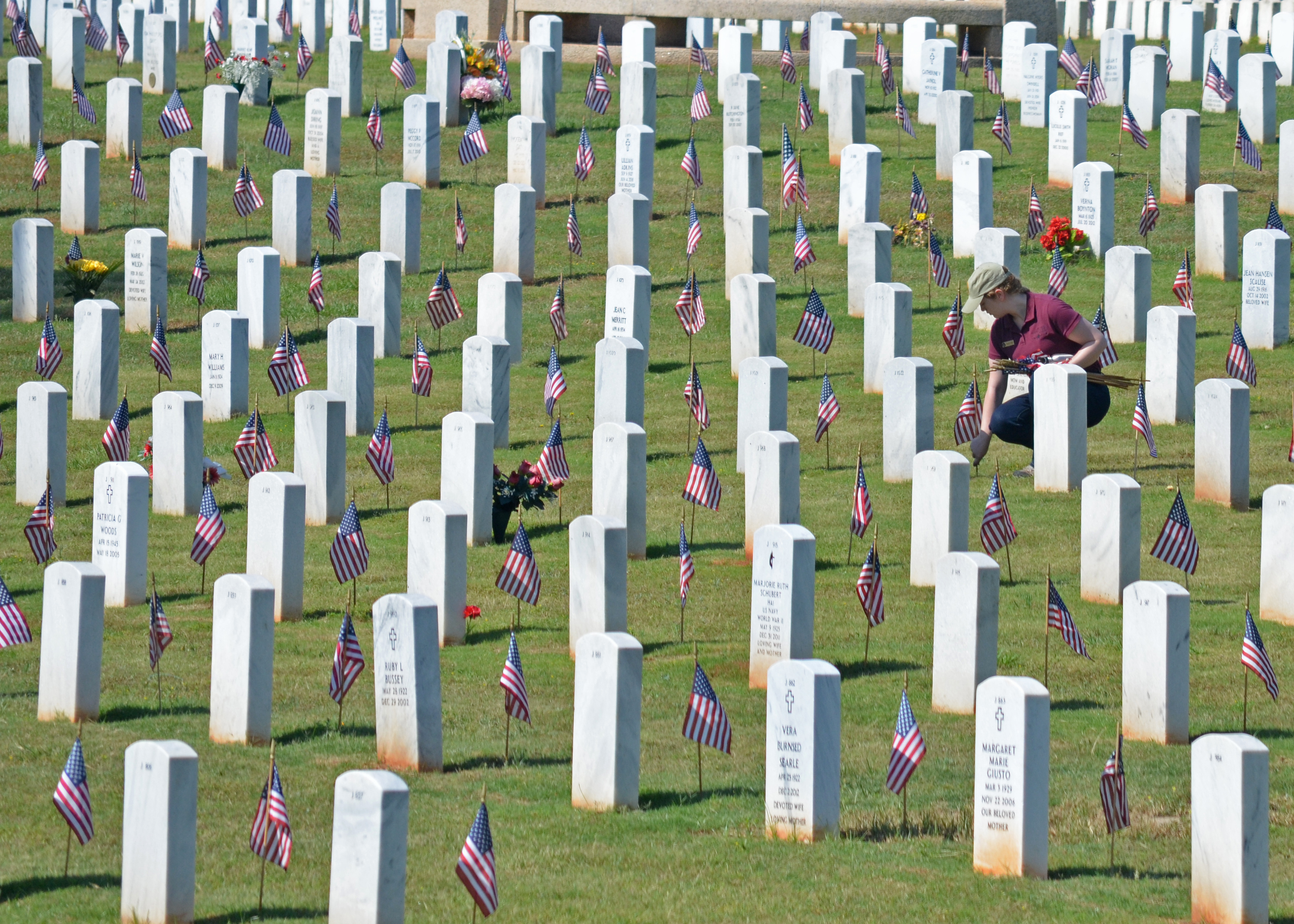 Rows of grave markers decorated with American flags. A woman crouches in front of grave stone and straightens a flag.