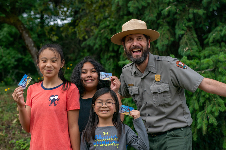 Three smiling girls show their Every Kid Outdoors pass standing next to a joyous park ranger.