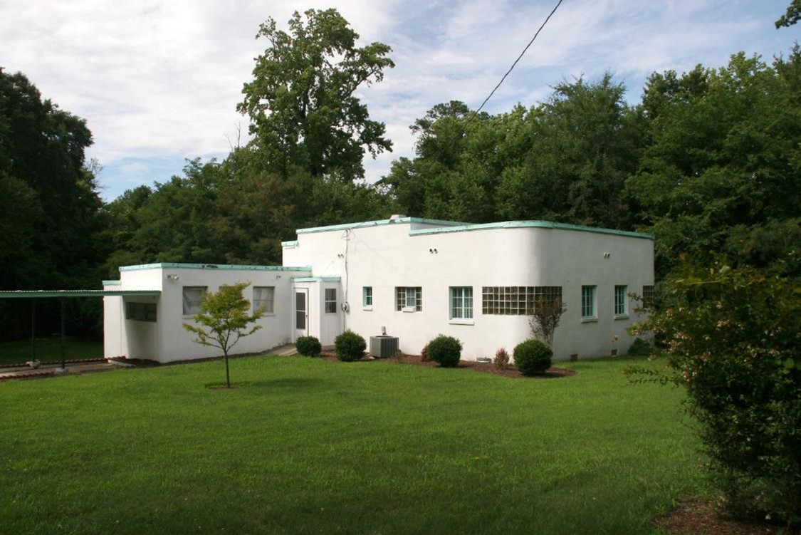 A white building on green grass in front of green trees and a blue sky with white clouds.