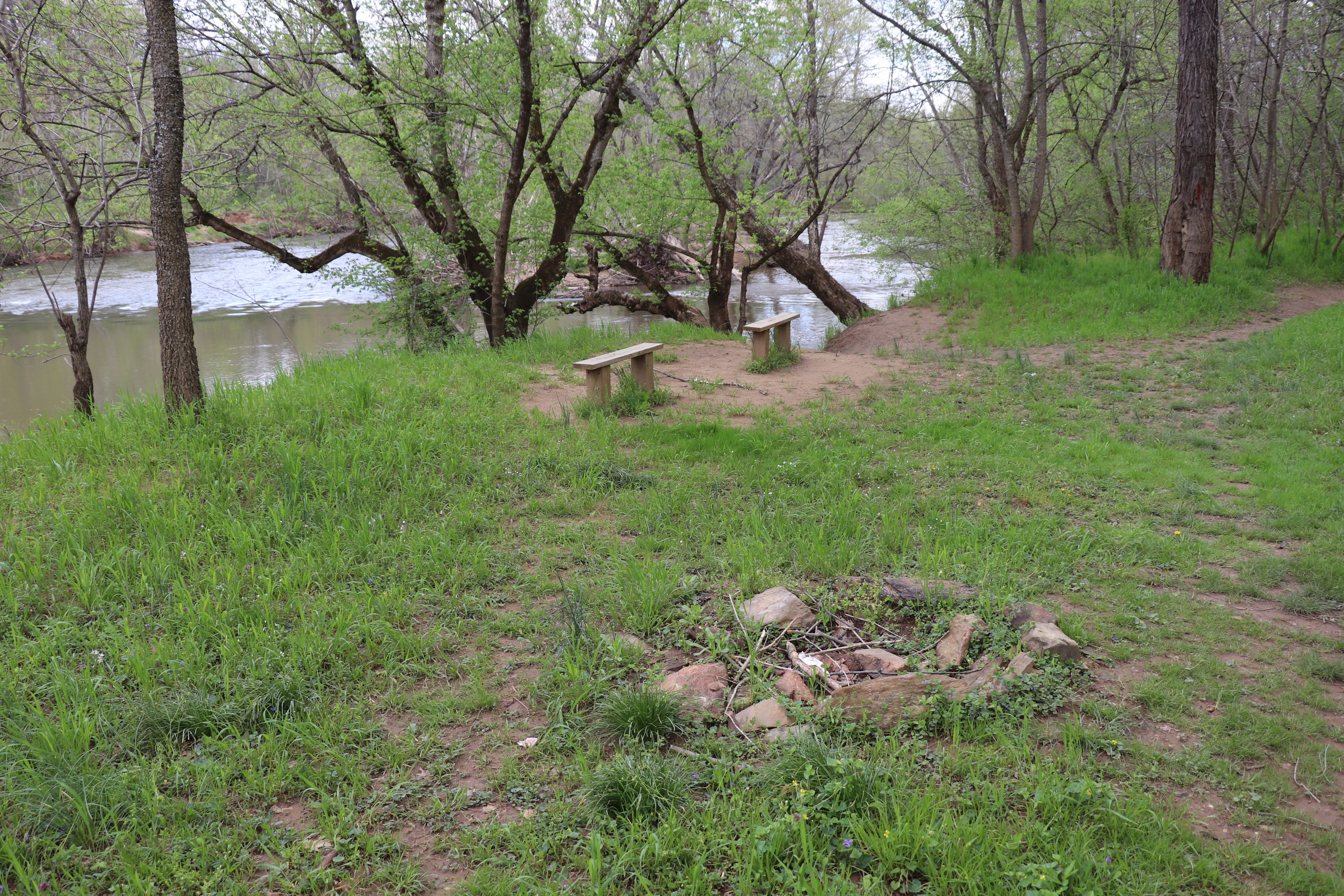 Two simple wooden benches face the river, at left, along a grassy trail through an open woodland that skirts a small circle of stones in the foreground.