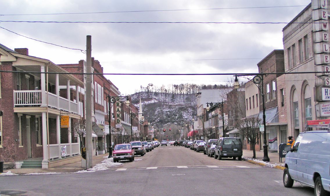 A street view of a small town, historic downtown with brick buildings, store fronts, lamposts lining the sidewalks, and cars parked on either sides of the street.