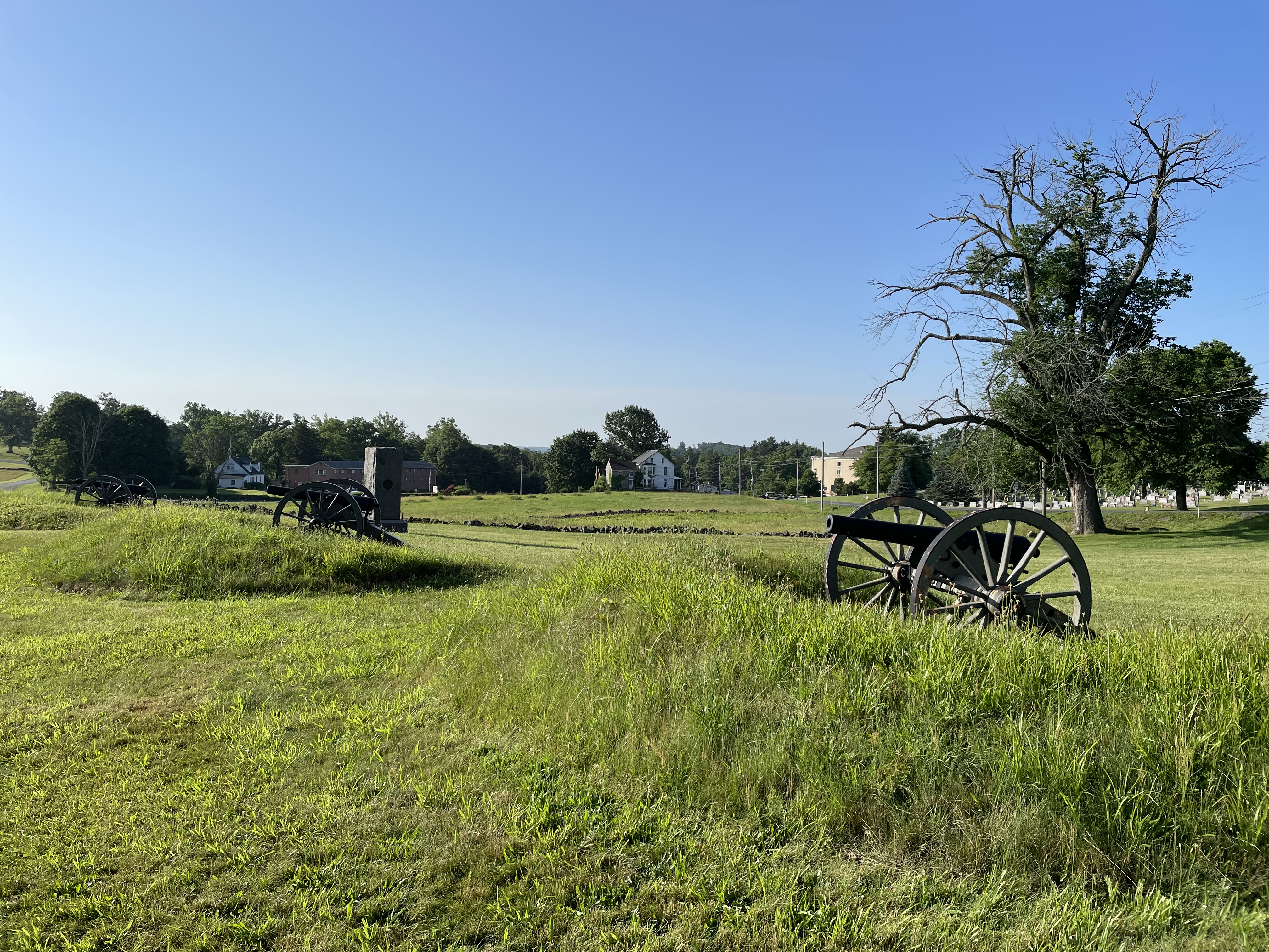 Canon and a granite monument in the foreground frame the view of the fields; a white house and a long brick building are at the horizon to the left and cemetery headstones are visible across the road at right.