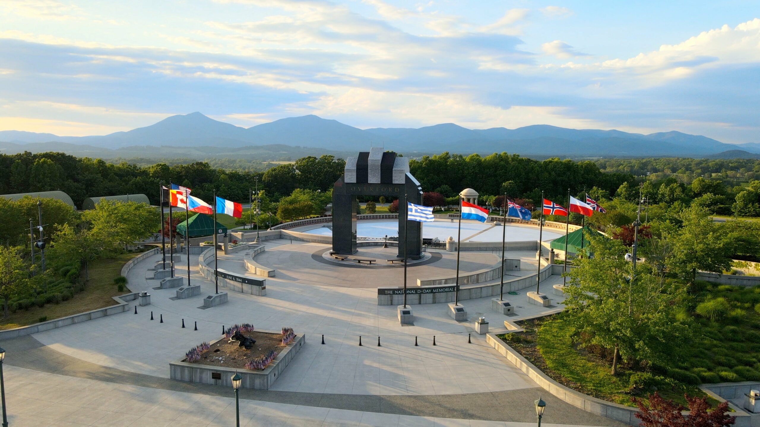 The National D-Day Memorial is seen from an elevated view, with blue sky, mountains, and trees surrounding it.