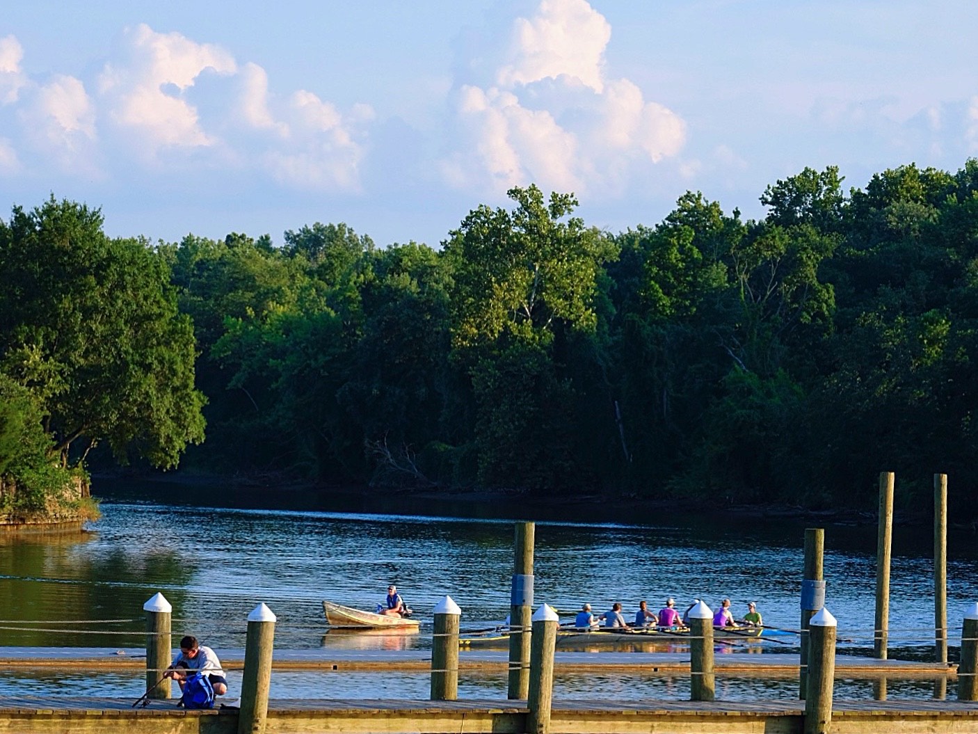 Bladensburg Waterfront Park A river landscape is surrounded by a forested edge and a blue sky with large, white clouds. A dock is in the foreground, with a man fishing off it. Behind the dock, people sit in a large rowboat.