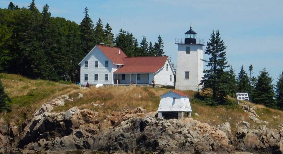 White building next to lighthouse on rocky ground with forest next to it.
