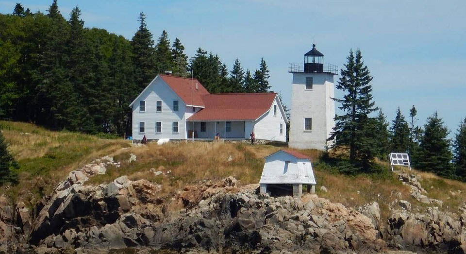 White building next to lighthouse on rocky ground with forest next to it.