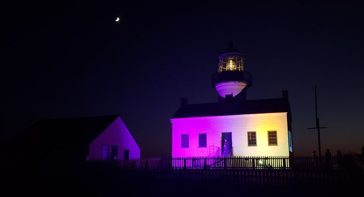 The lighthouse at Cabrillo National Monument is illuminated in purple and gold against a dark night sky a white building with a lighthouse is illuminated in purple and gold