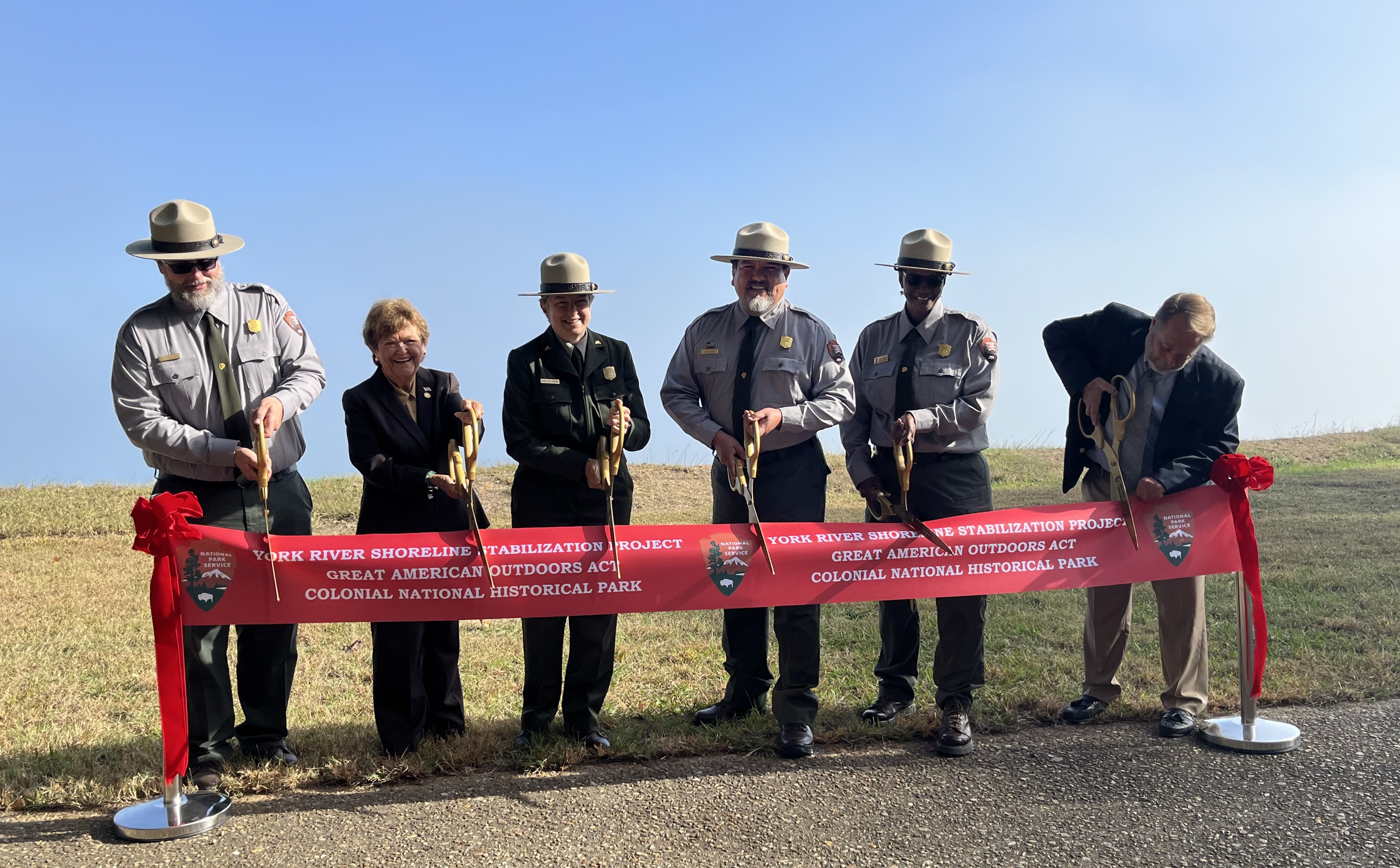 COLO ribbon cutting Six people, including four wearing National Park Service uniforms and two in business suits, pose with giant scissors to cut a red ribbon that says York River Shoreline Stabilization Project, Great American Outdoors Act, Colonial National Historical Park