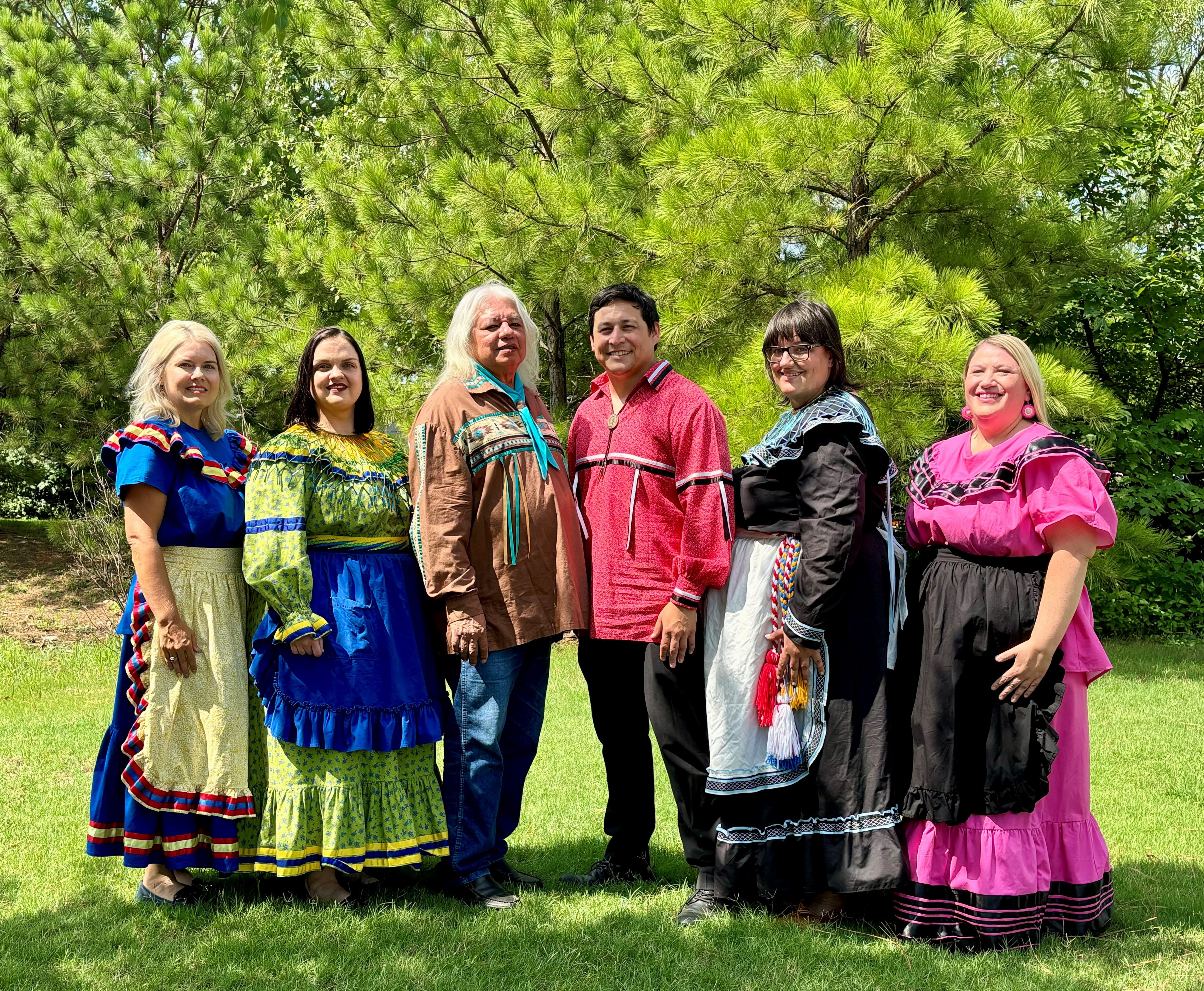 Chickasaw Nation Reburial Team 7.18.24 Six people from the Chickasaw Nation pose in Tribal attire in front of a grove of pine trees.