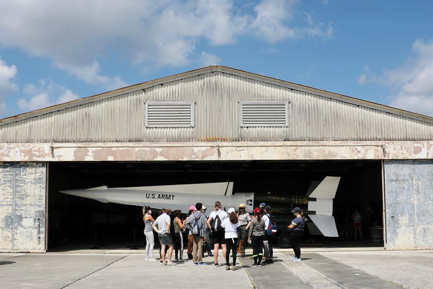 Cold War Hangar Several people face away, looking at a low hangar; inside is a white plane labeled "U.S. Army".