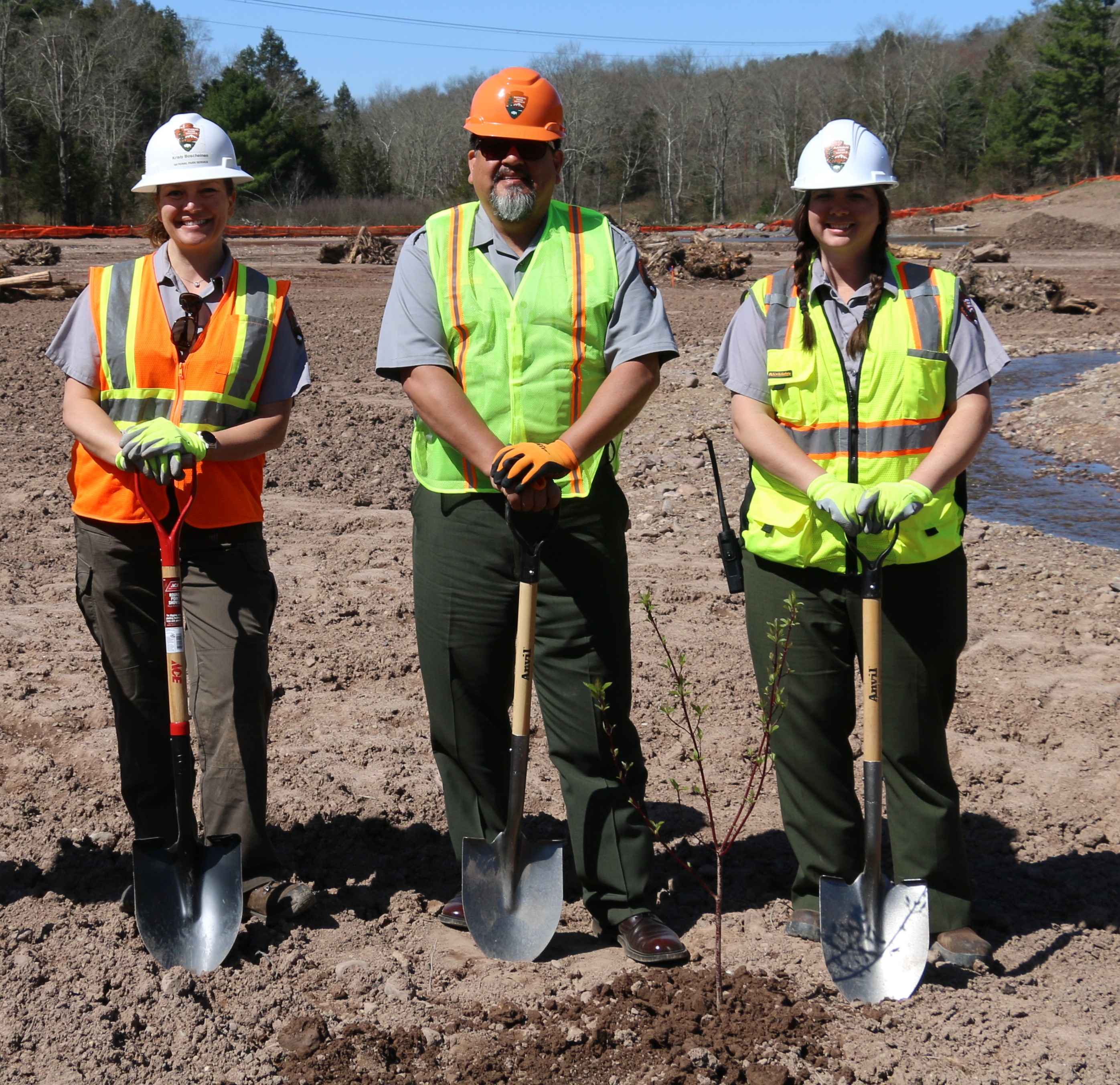 Three people wearing hard hats and holding shovels stand behind a tree sapling they just planted.