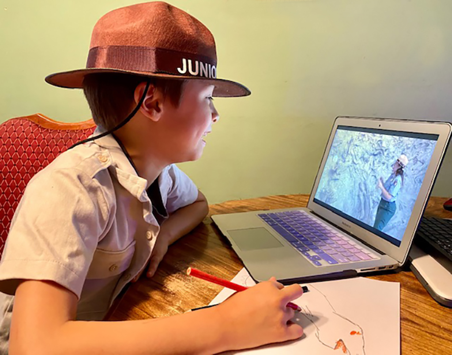 DINO_Junior Ranger_virtual A junior ranger in uniform with a hat sits at a table, looking at a laptop showing an image of an NPS park ranger in the field.