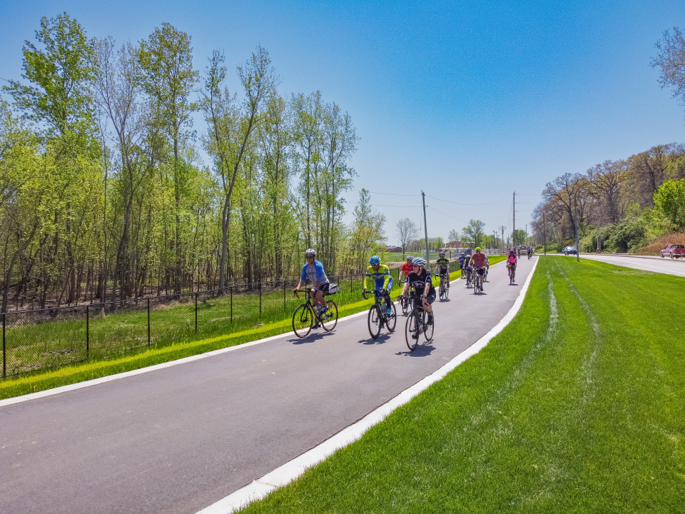 Old Chain of Rocks Bridge Park (St. Louis, MO) A group of adults clad in bright colors of blue, red, purple and yellow ride bikes along a black asphalt road, surrounded by green grass and towering trees underneath a bright blue sky