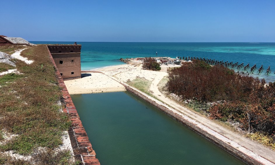 View looking down along brick fort wall at water-filled moat with ocean in background