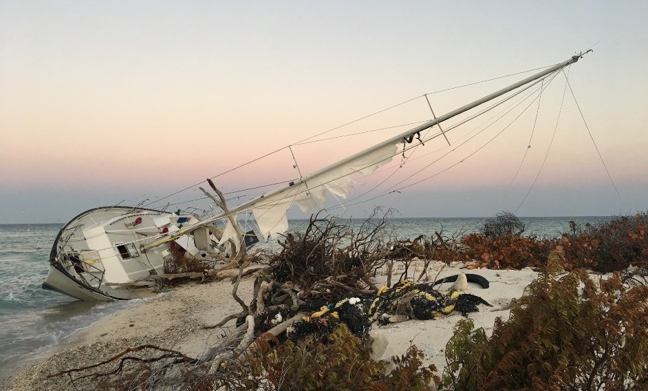 A single-masted sailboat lies on its side along a sandy shoreline