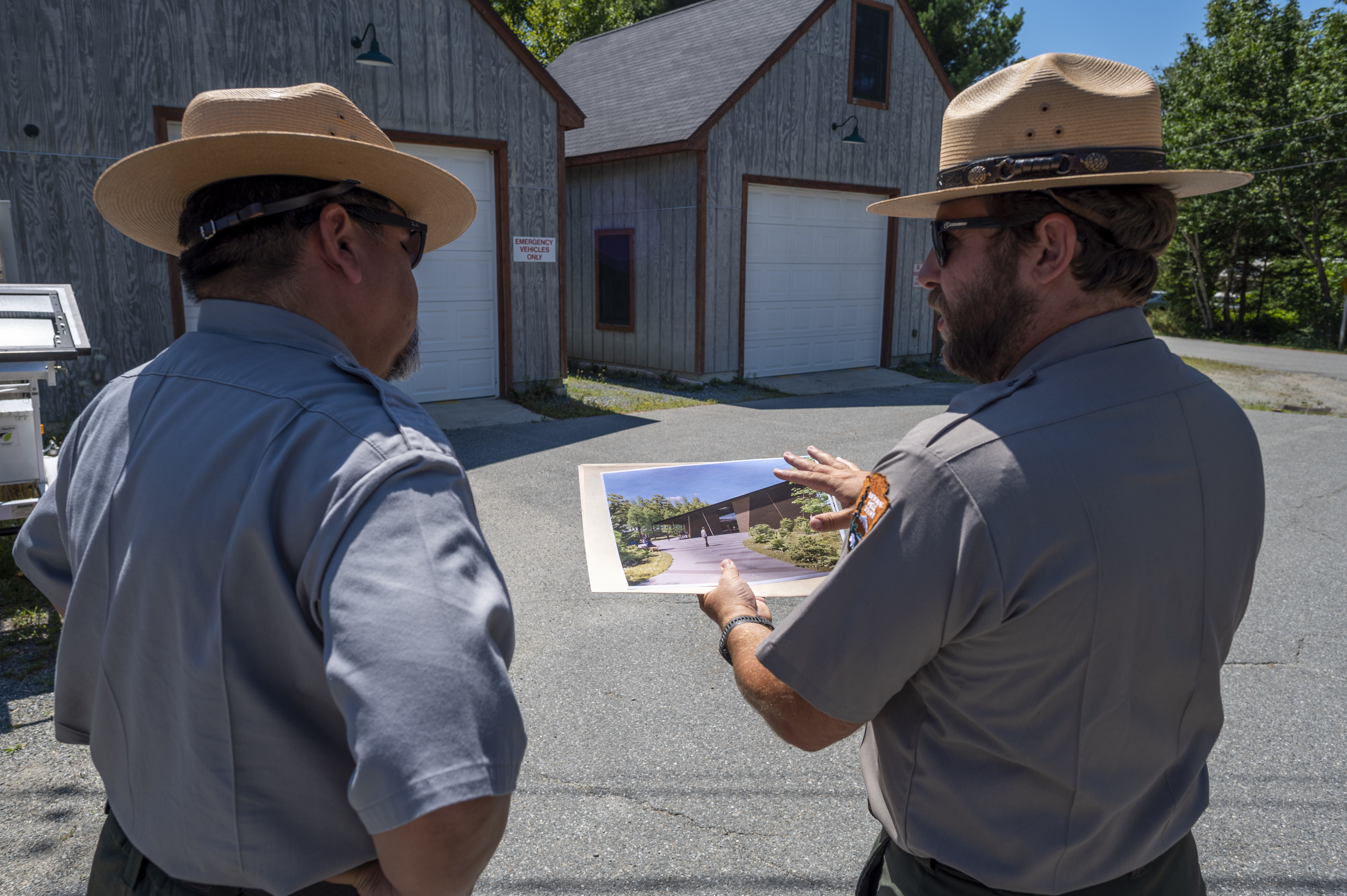 Against the backdrop of two outbuildings, two men face away, both wearing flat hats and gray uniform shirts.  The man on the right is holding an image of a different building.