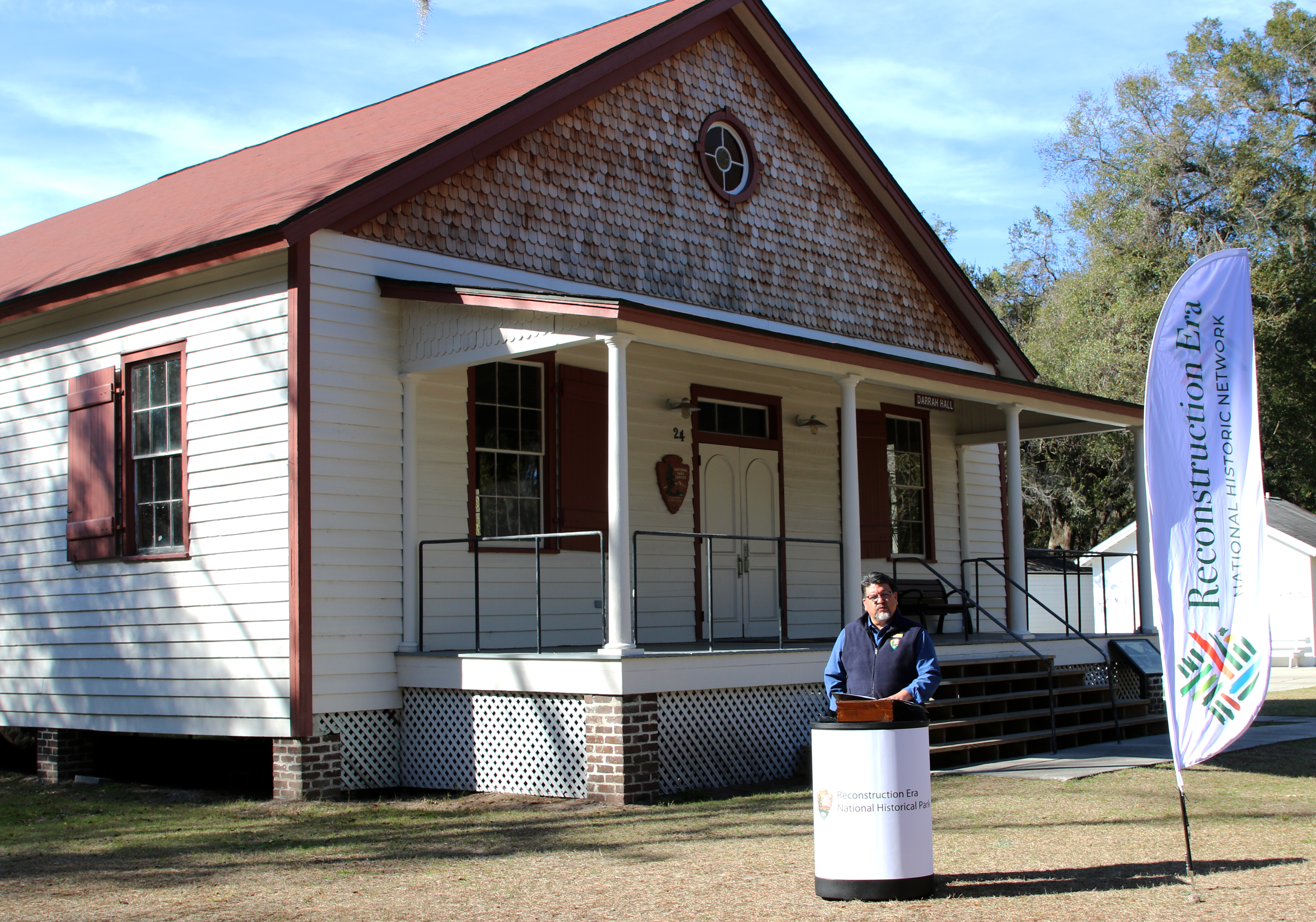 Director Sams stands in front of a long white building with red trim, a large front porch and wood shingled soffit.