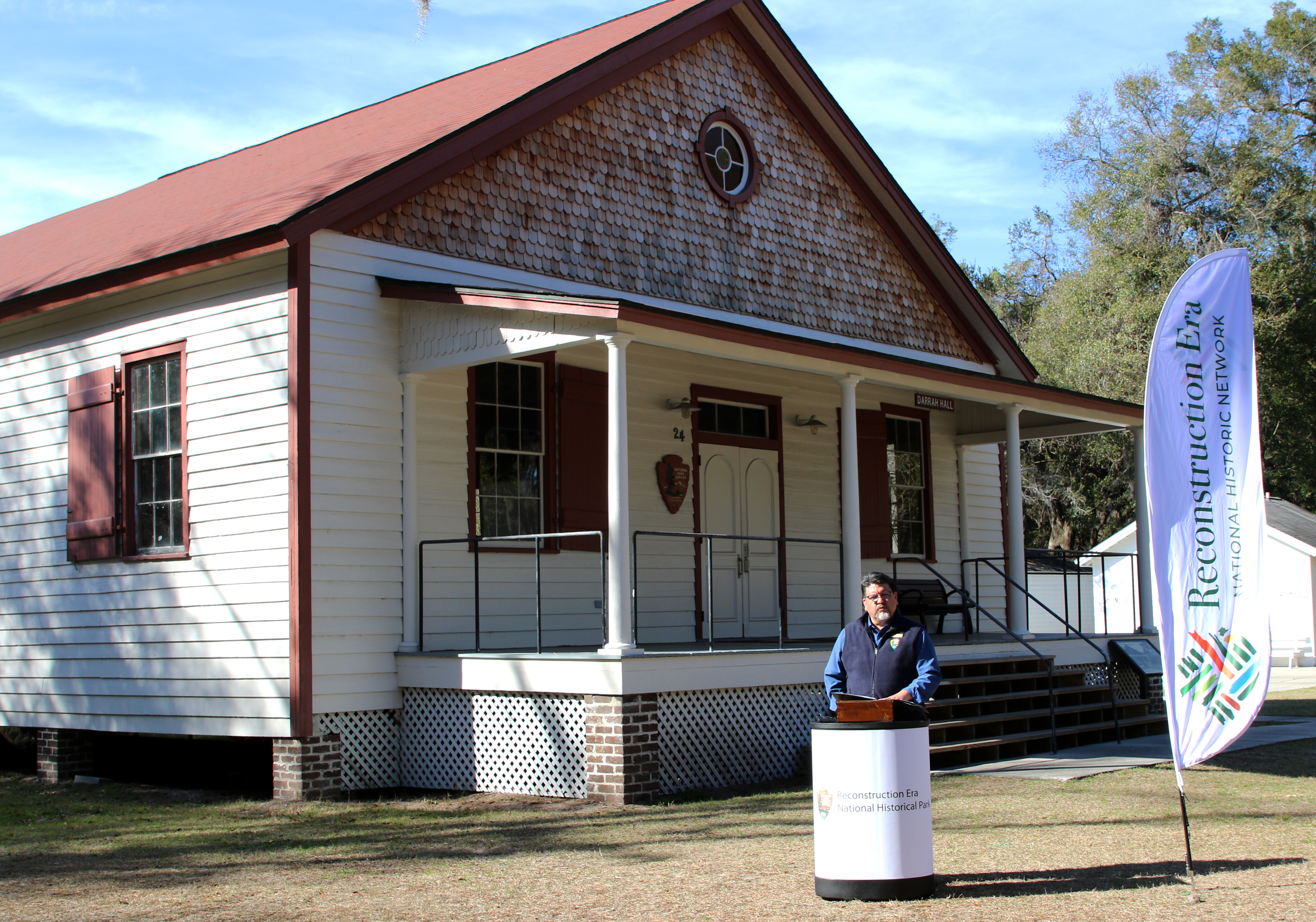 Director Sams at REER Director Sams stands in front of a long white building with red trim, a large front porch and wood shingled soffit.