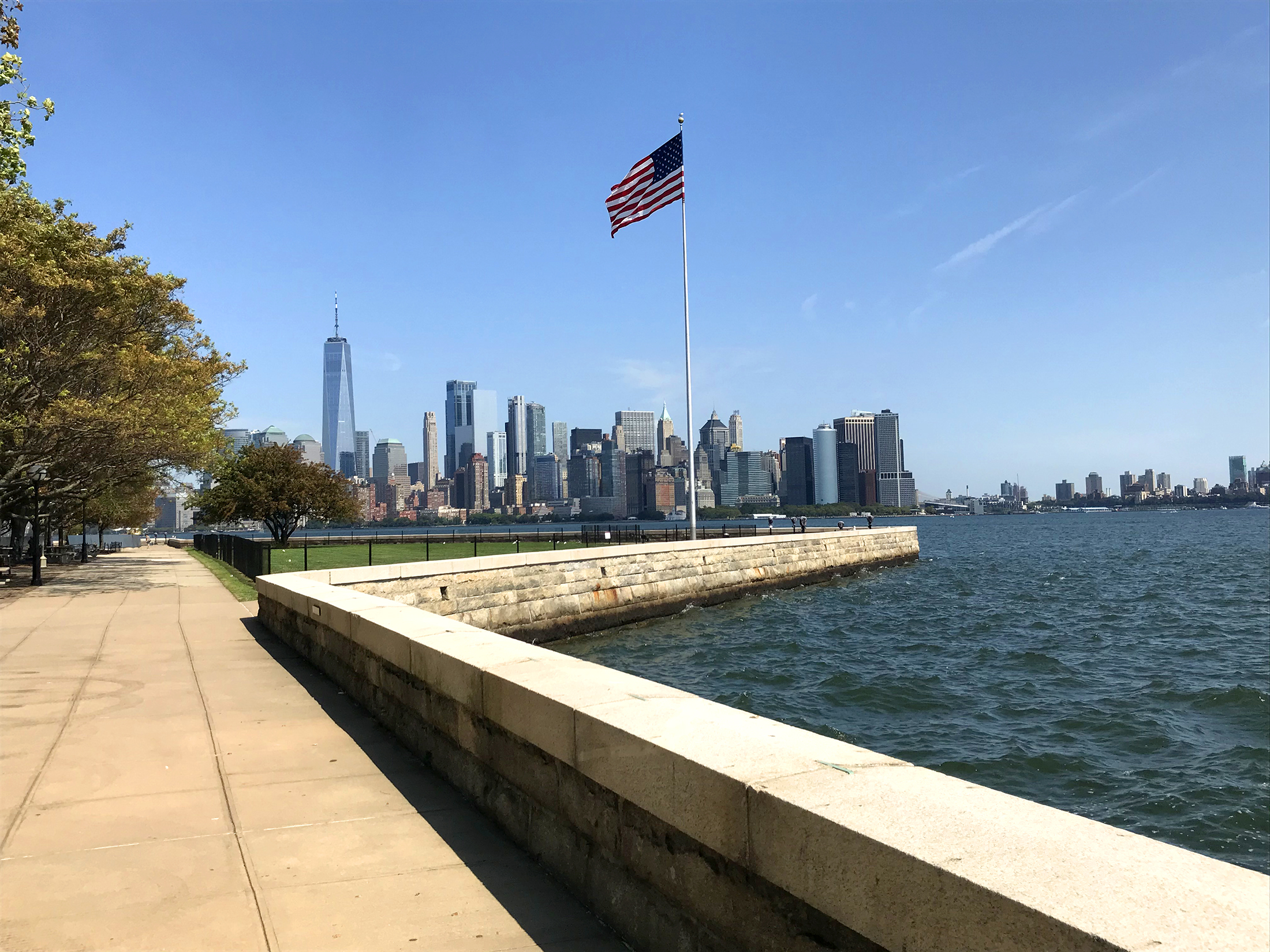Ellis Seawall A low stone wall along a body of water with an American flag on a flagpole near the wall. Tall skyscrapers in the distance.