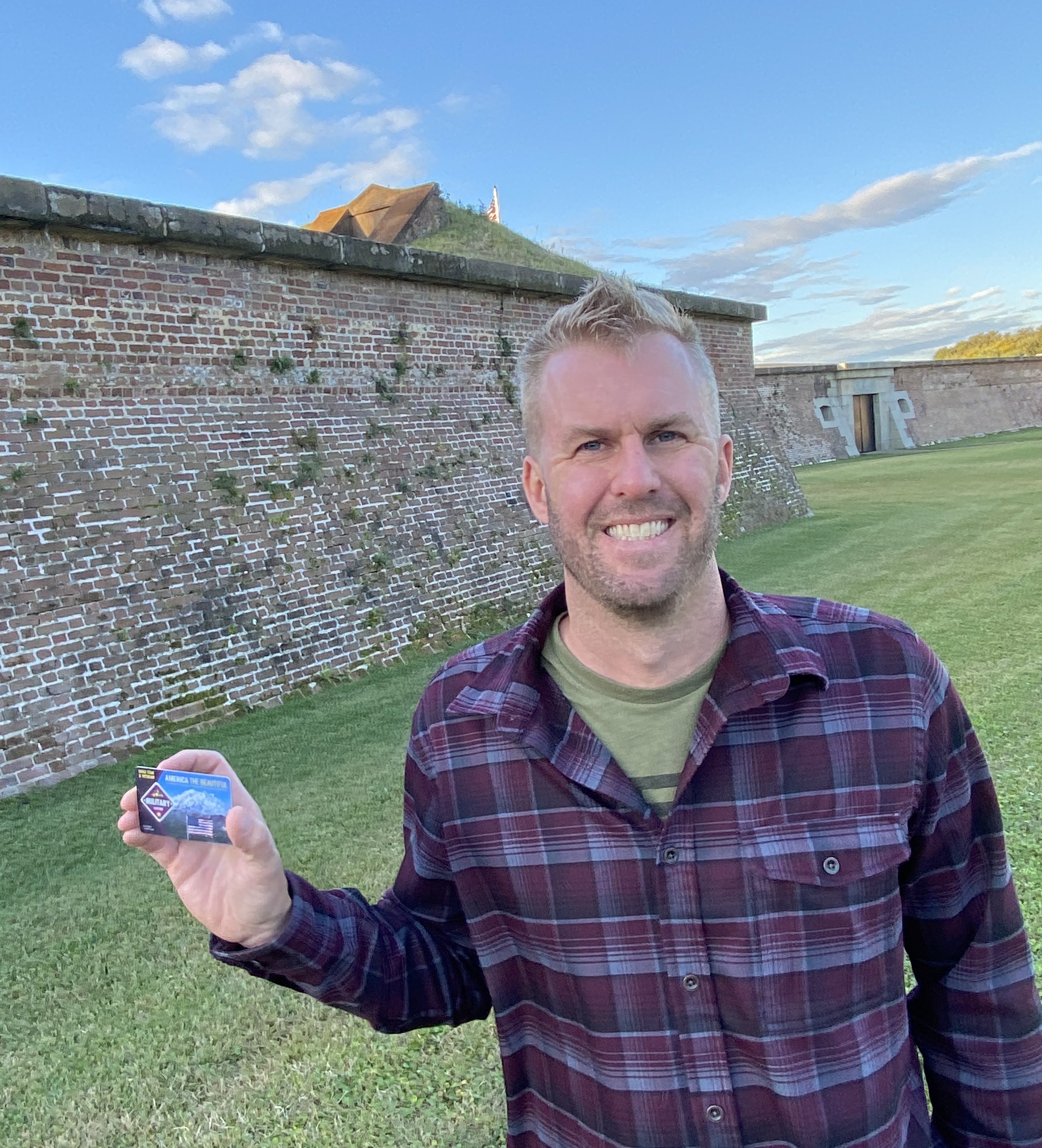 A young man wearing a plaid shirt holds a park pass in his right hand. He is standing on grass in front of the wall of a historic fort. A young man wearing a plaid shirt holds a park pass in his right hand. He is standing on grass in front of the wall of a historic fort.
