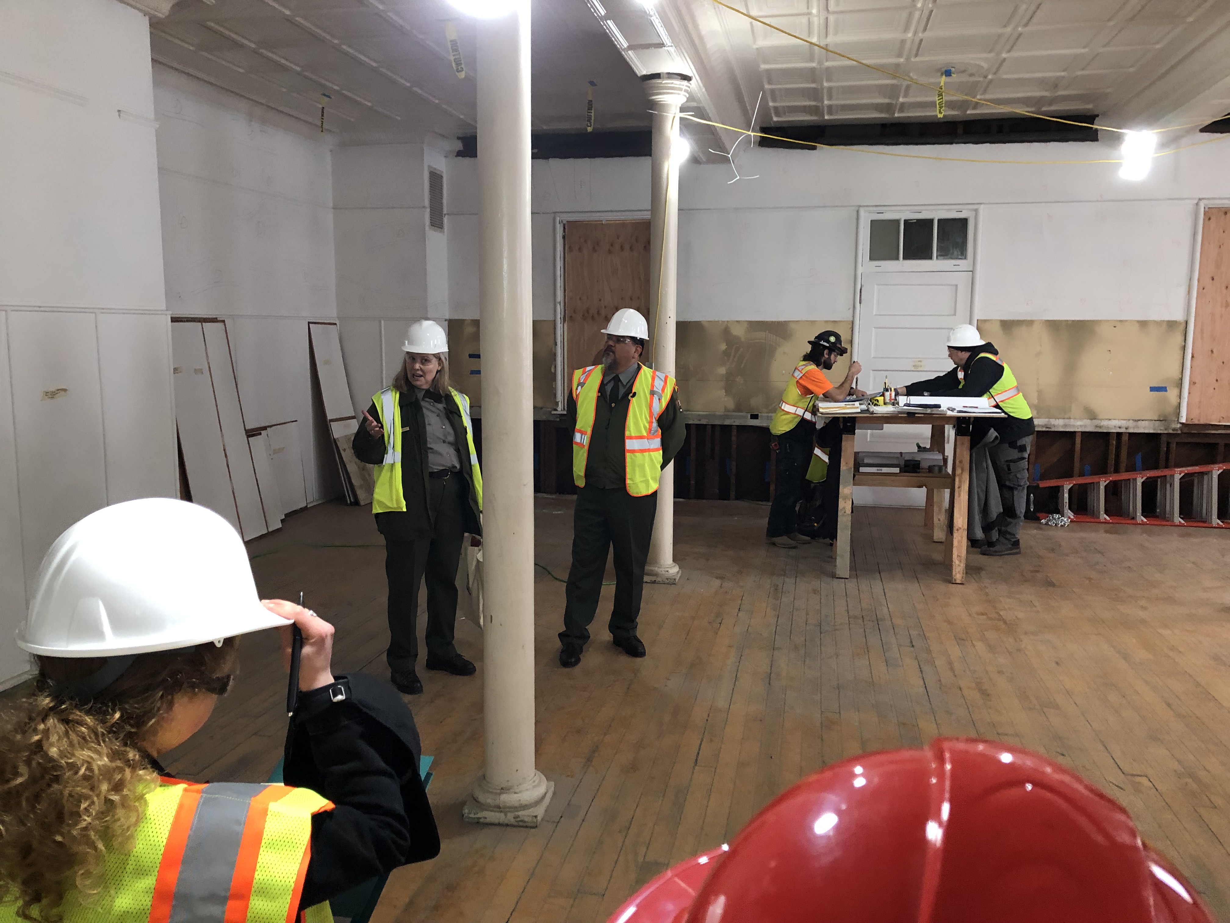 Six individuals stand around a room, four facing each other in conversation and two are standing at opposite ends of a table towards the back of the room. All are wearing hard hats, googles, and reflective vests.