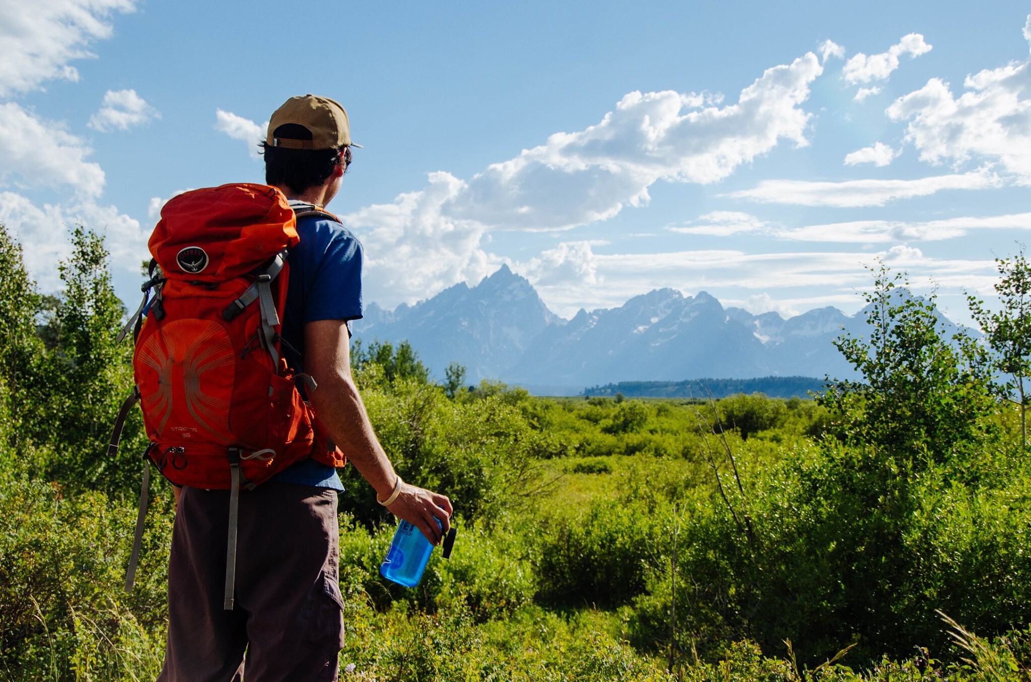 Start planning your 2025 national park visit A hiker looks out on Teton Range holding a water bottle and wearing a backpack.