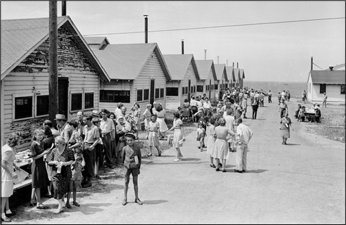 Refugees gather Fort Ontario Barracks In black and white. On the left, a line of people dressed in 1940s-era clothing stand in front of eight cabins. The cabins are along a dirt road where many more people are gathering.