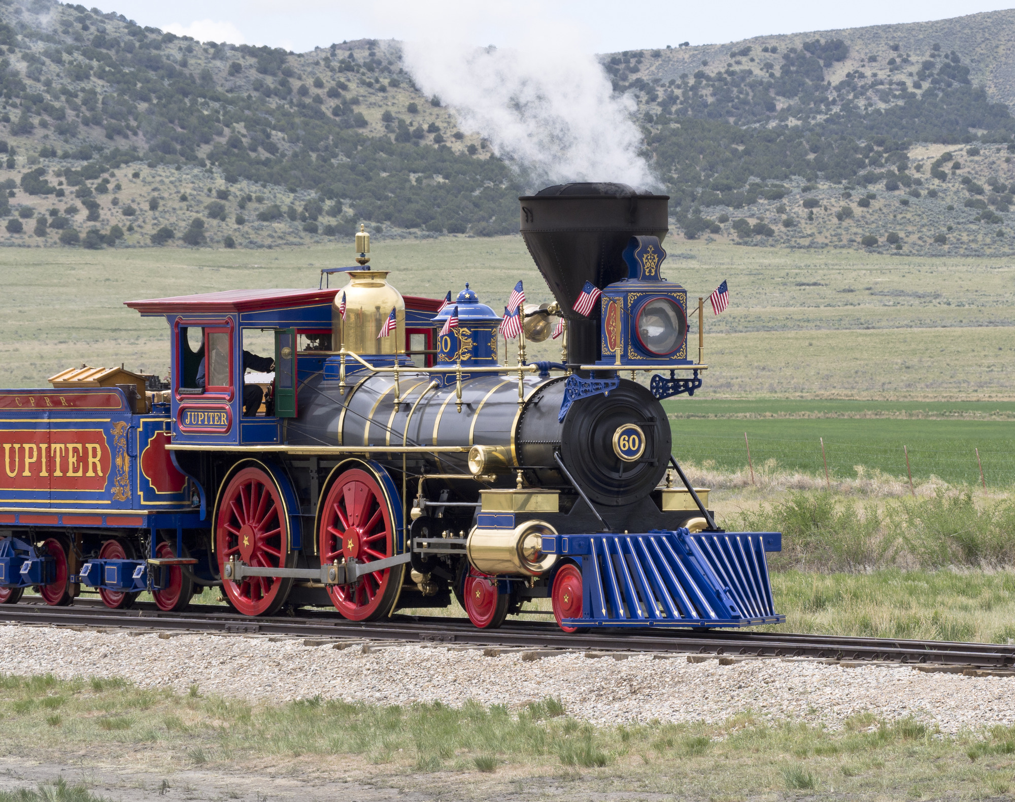 A red, blue, and black steam locomotive decorated with small American flags waving in the wind, moving forward on a railroad in a sparse landscape with hills in the distance. "Jupiter" on the side of the locomotive.