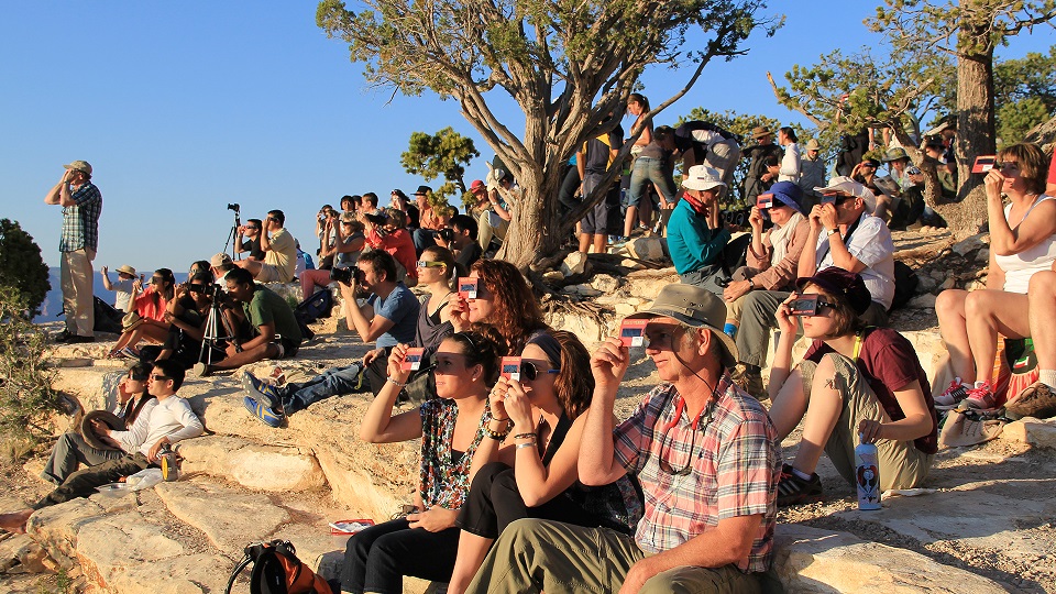 Dozens of people sit or stand outside on a rocky slope and all face the same direction (left) while holding card shaped solar viewers or while wearing solar eclipse glasses. It is a sunny day with a blue sky and trees in the background. 