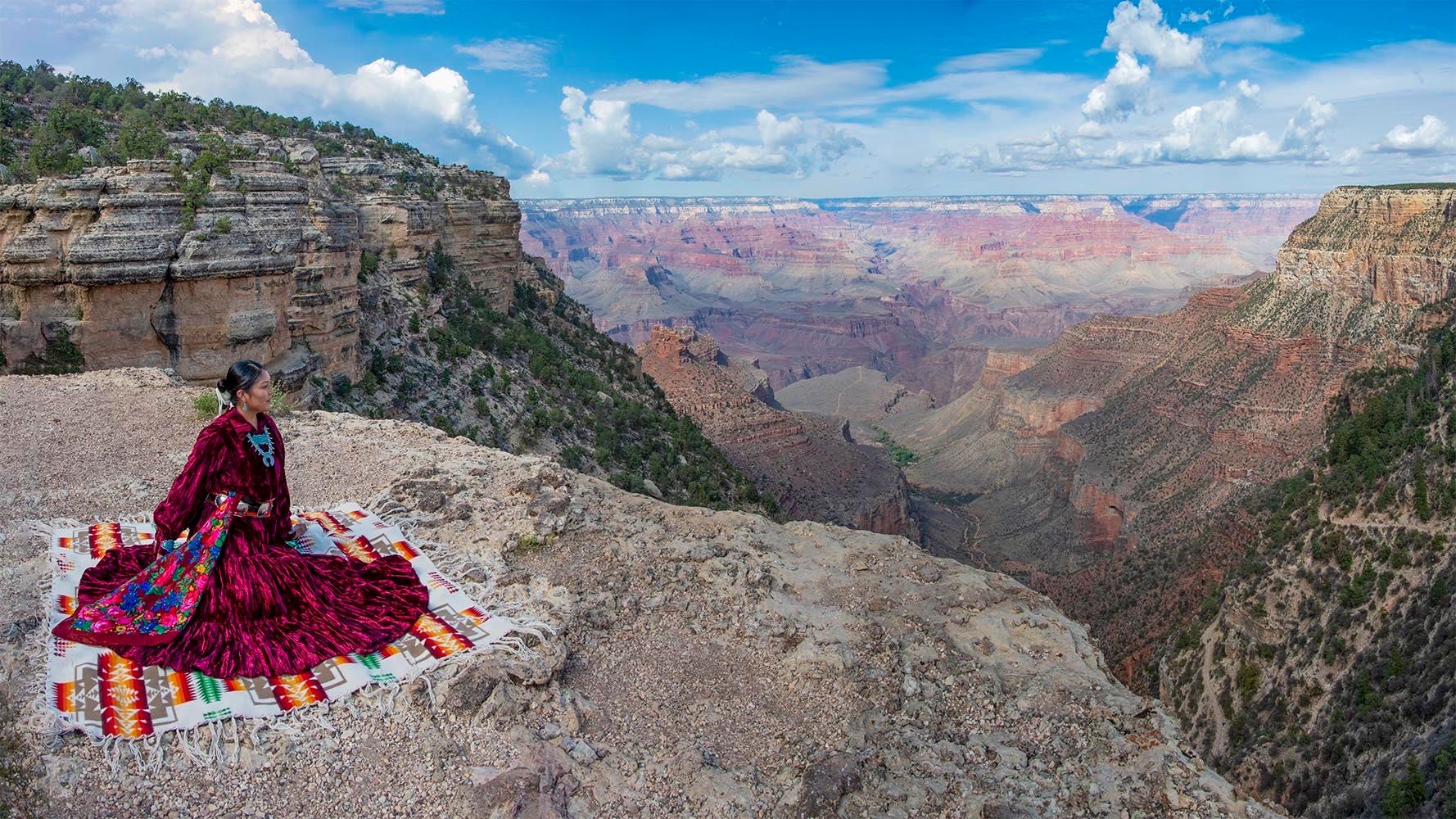 A Native American woman looking over the Grand Canyon. Native American woman in beautiful traditional long red dress sitting on a blanket and looking over the Grand Canyon.