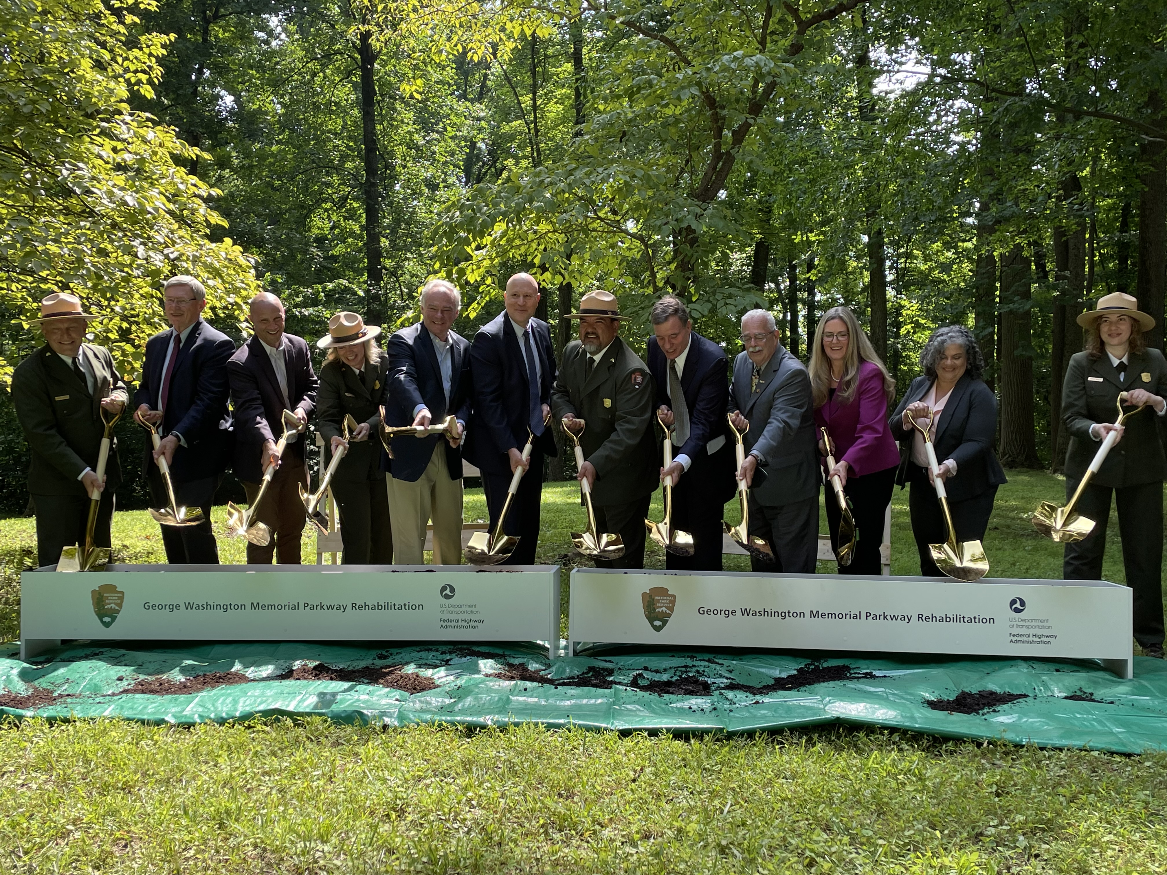 Various people, wearing suits and green uniforms, dig dirt behind a banner reading "George Washington Memorial Parkway Rehabilitation".