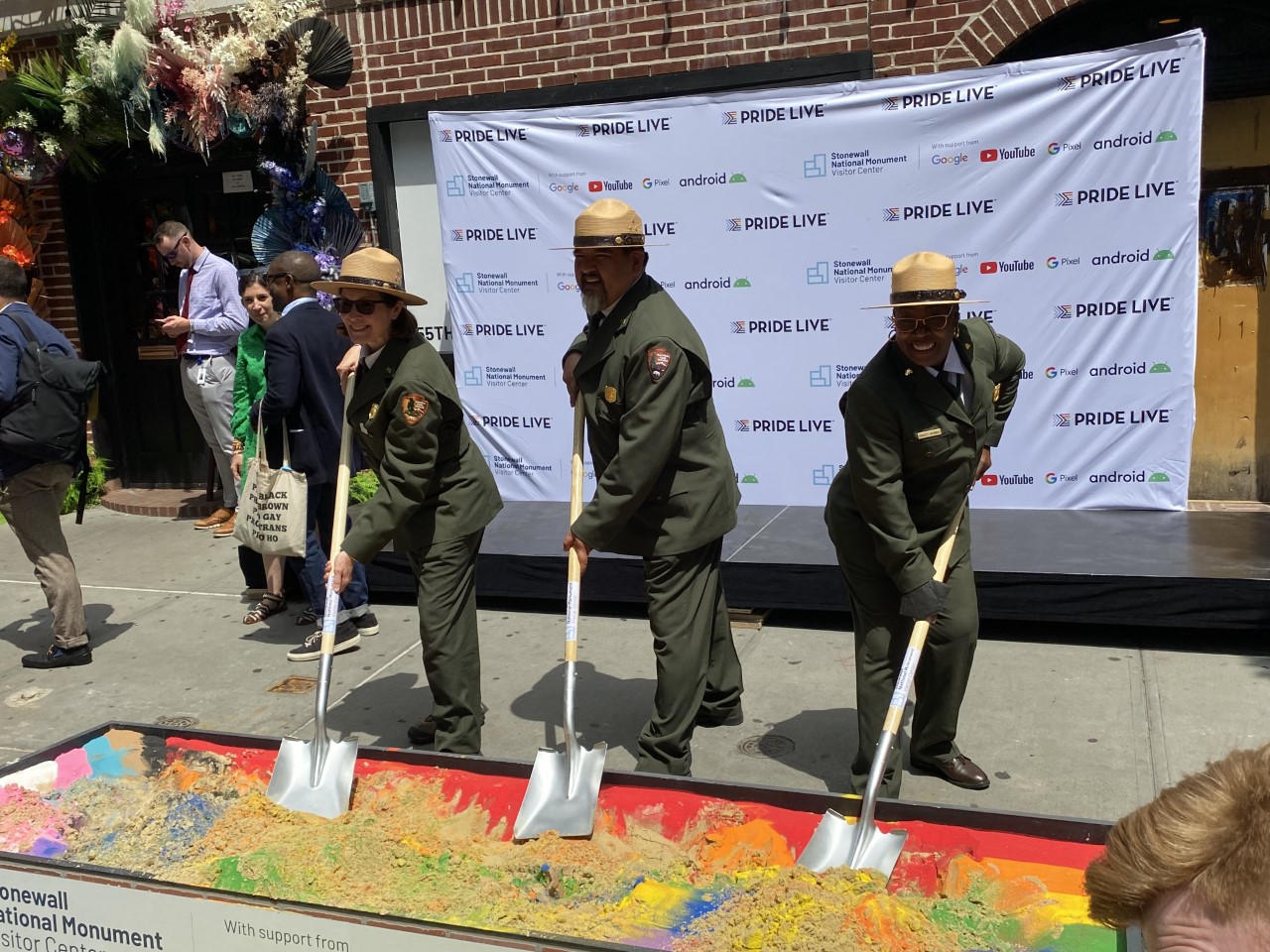 Ground Breaking STON June 242022 Three park rangers in green uniforms plant shovels into rainbow-colored dirt in front of a backdrop reading "Pride Live".
