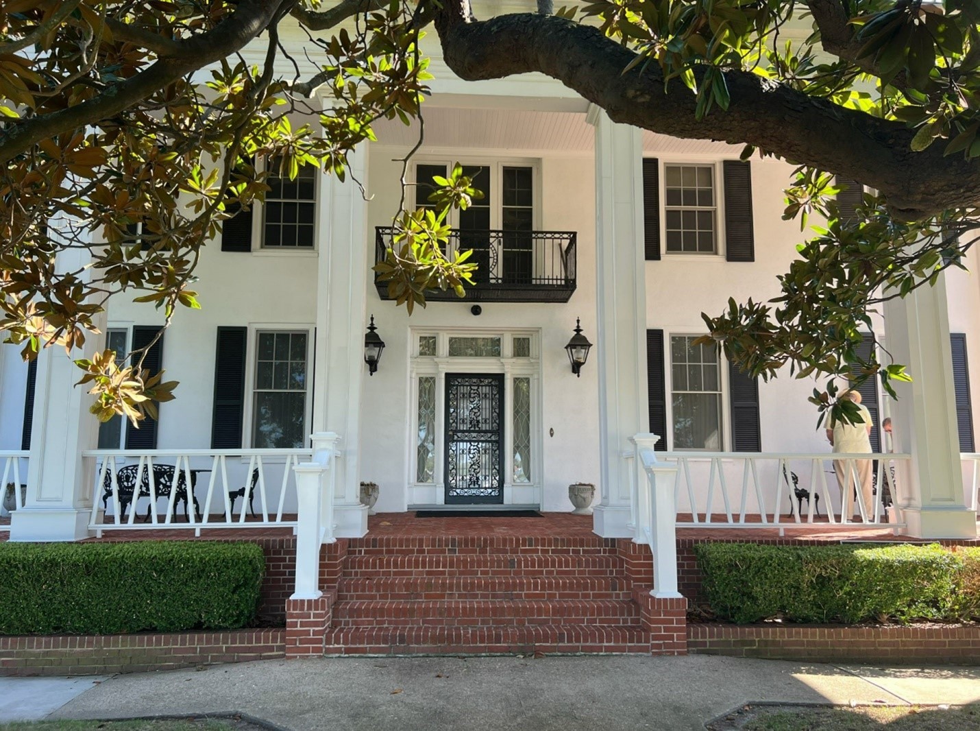Large, three-story white building with pillars and a front porch in the background and a leafy tree branch in the foreground.