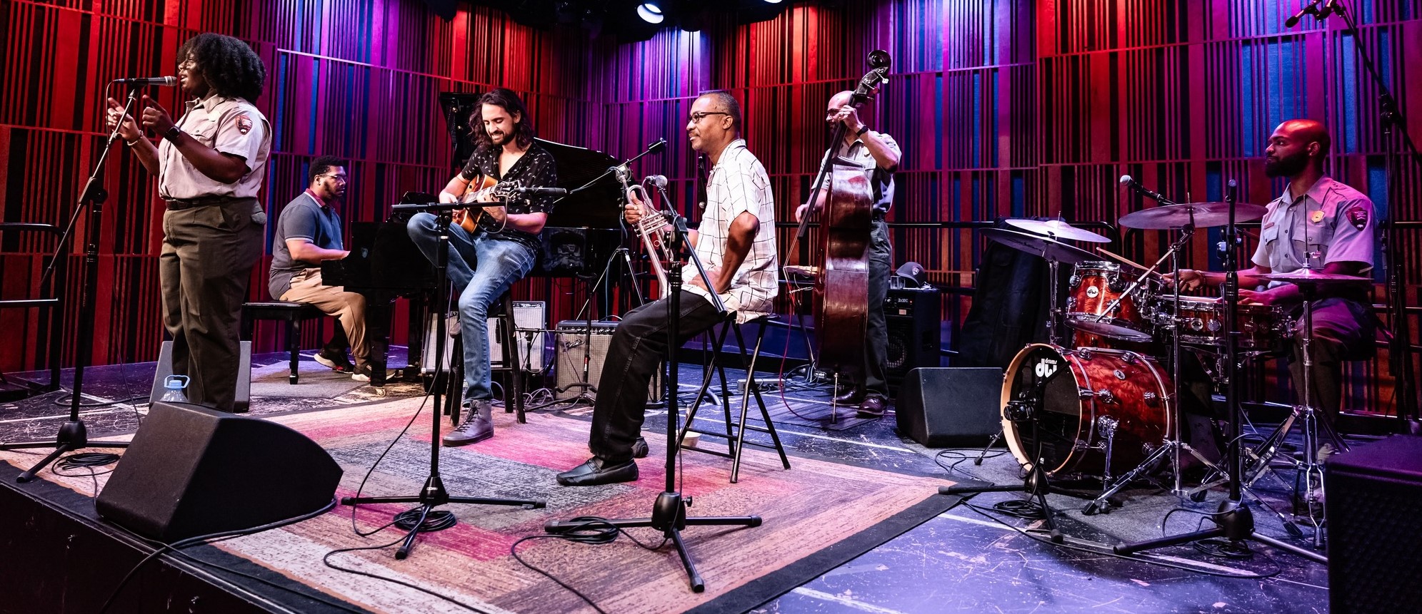 Musicians on stage. There are six musicians on a stage, including a vocalist, guitar player, piano player, a trumpet player, a drummer, and a stand-up bass player.
